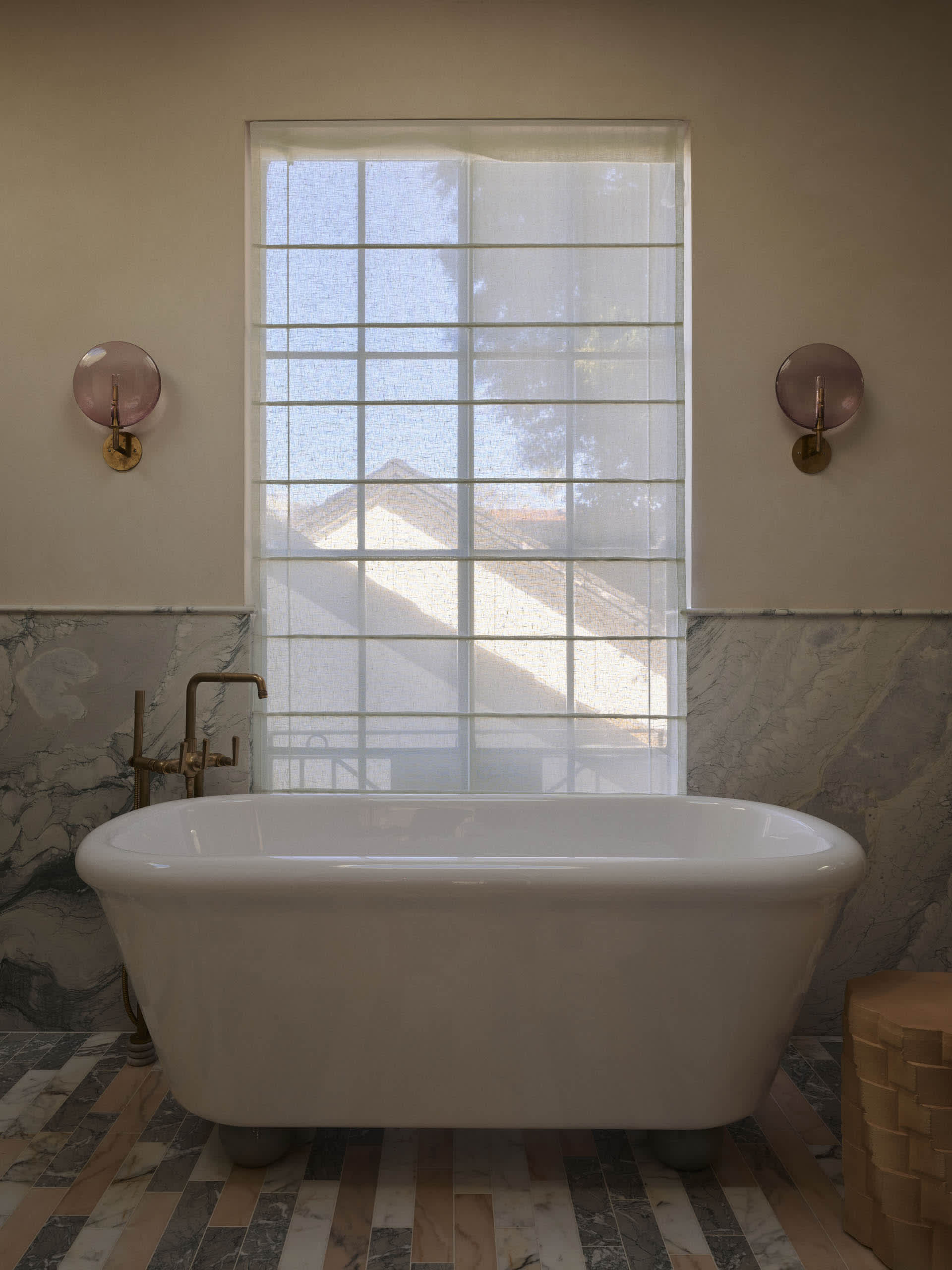 Bathroom with a large white bathtub under a window, surrounded by marble-like surfaces and white curtains, lit by natural sunlight