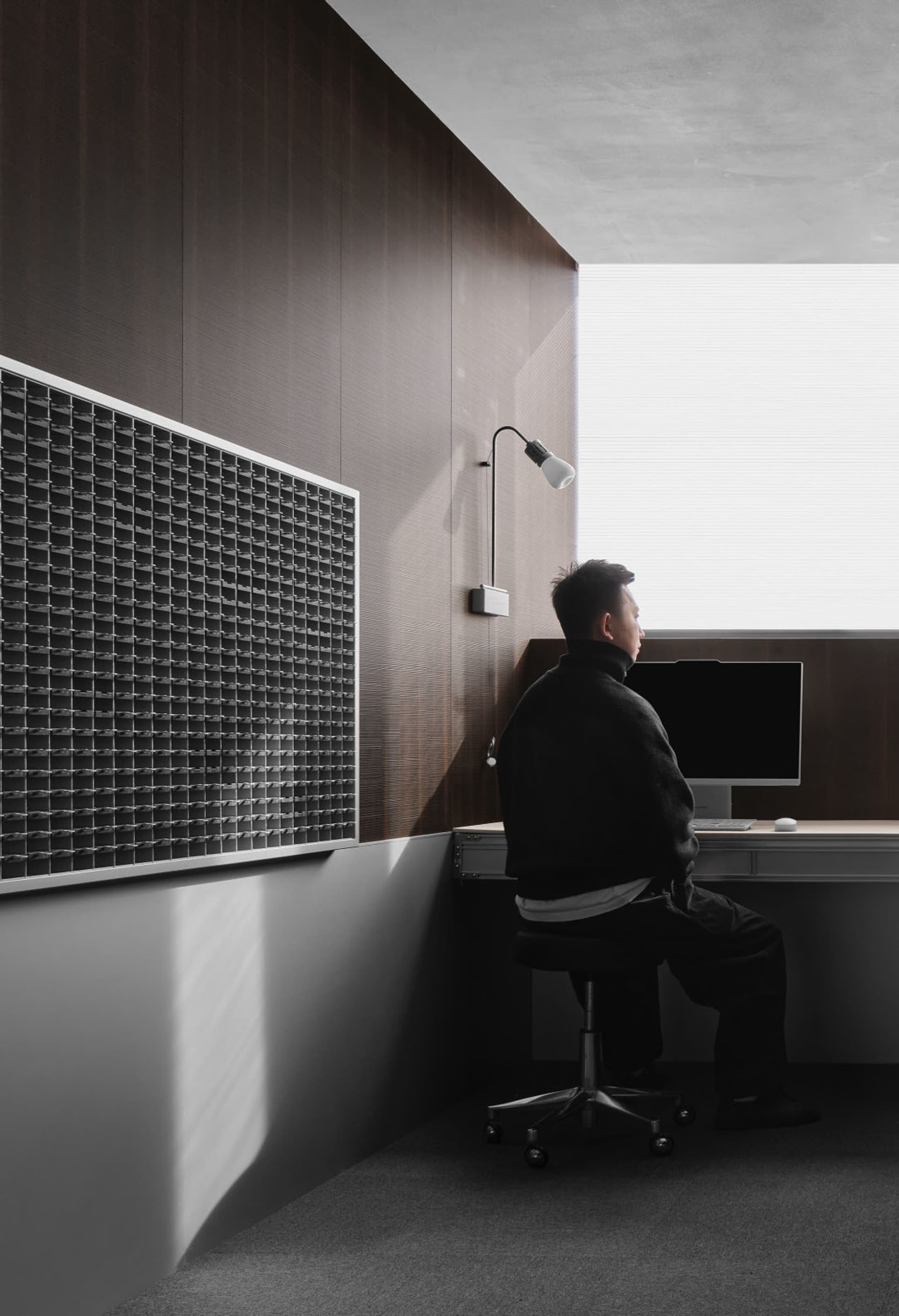 A man sits at a desk in a small office space with a wooden floor and a window, working on a computer