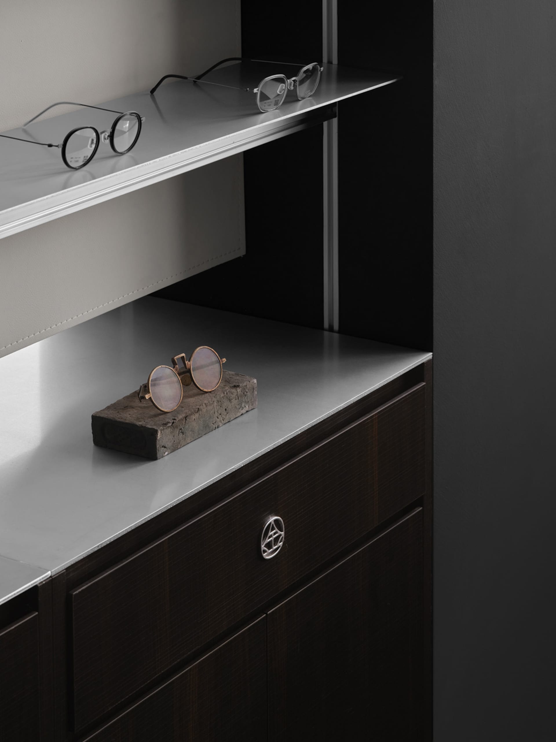 A dimly lit modern bedroom with a metal countertop, a bed in the background, and a pair of sunglasses and a small rock on the countertop