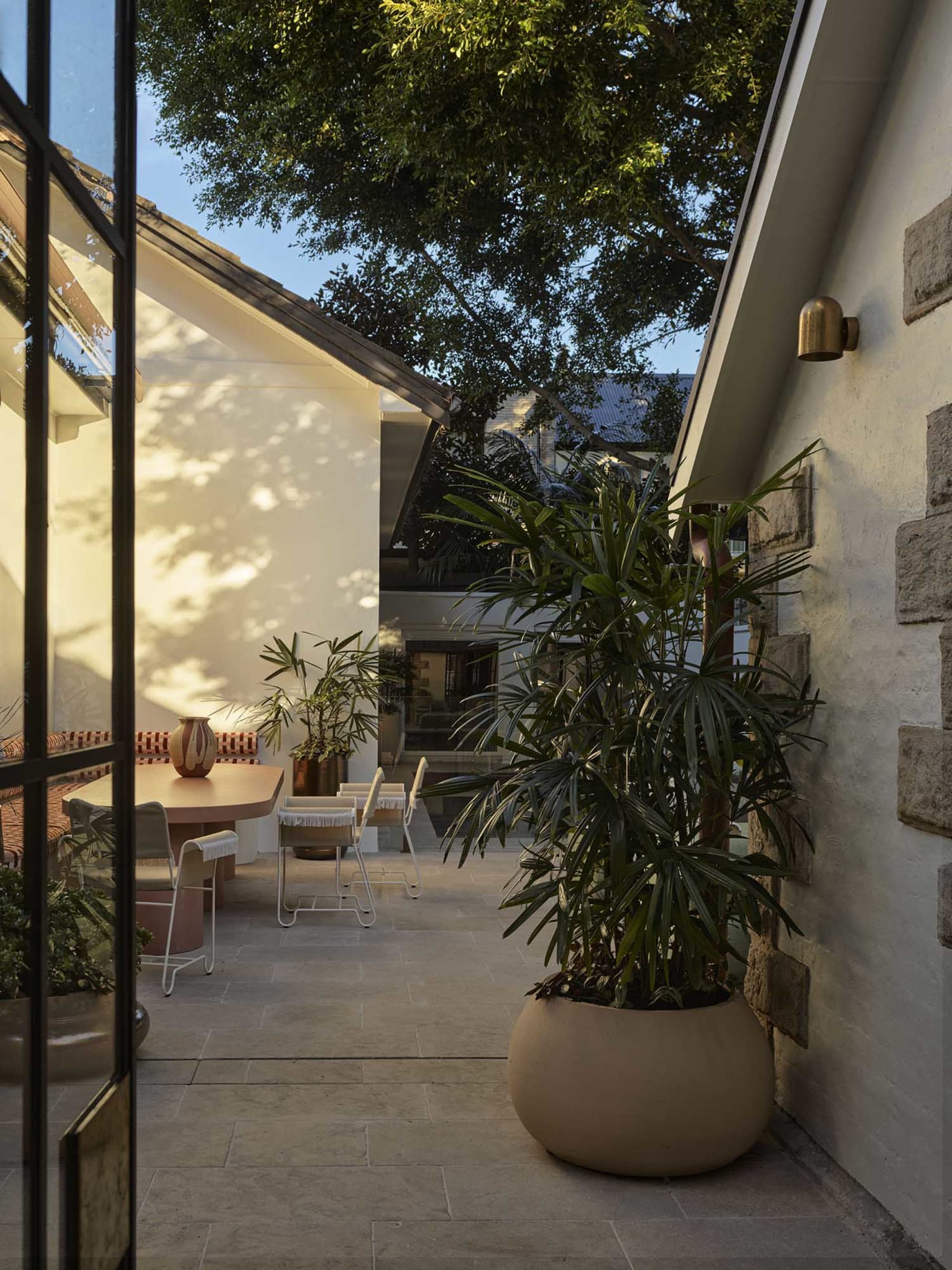 A residential patio with a dining table and chairs, surrounded by a potted plant, with a house in the background, featuring a mix of wood and stone elements, and natural sunlight creating a warm atmosphere