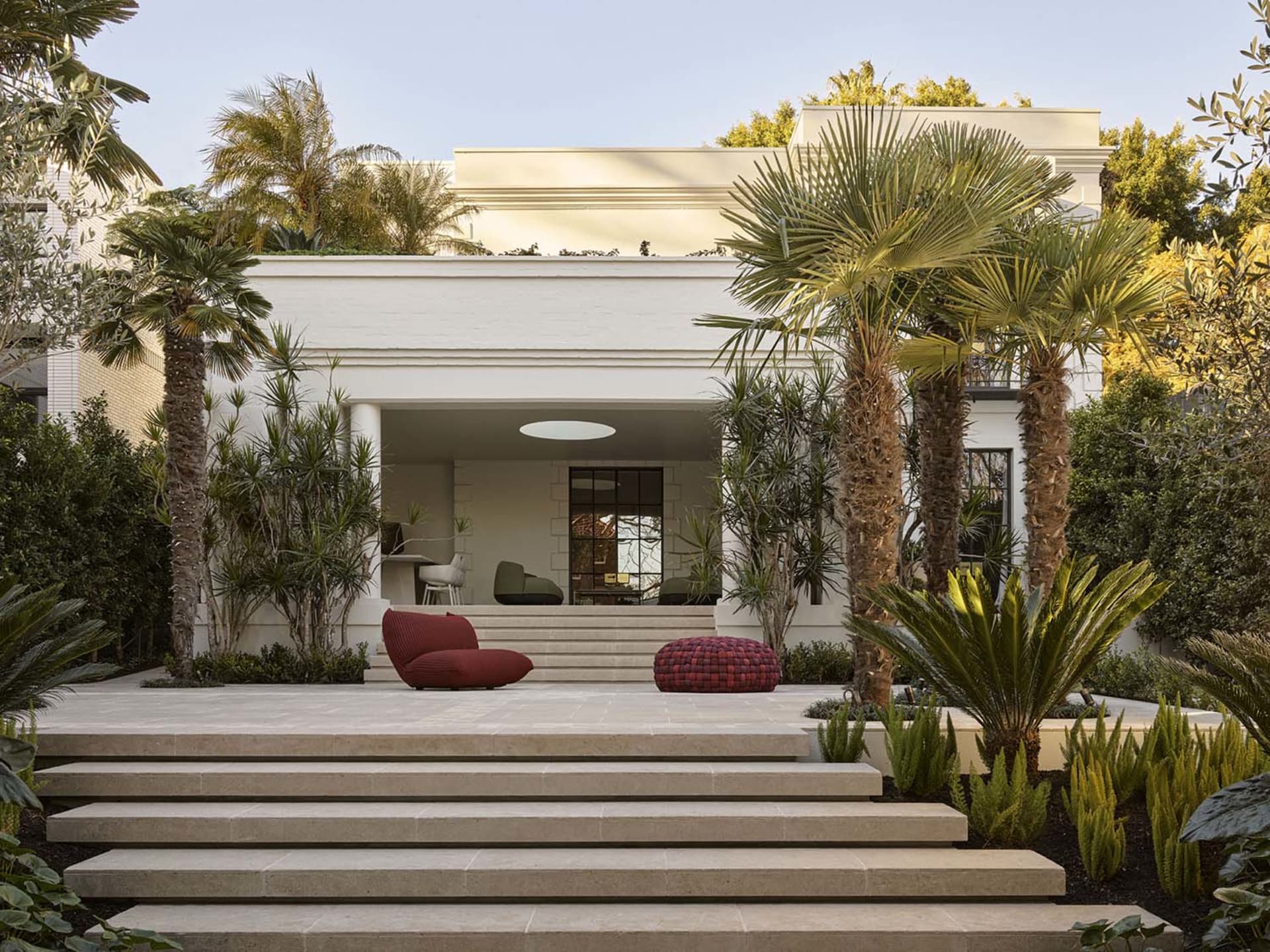 A modern house with a white exterior, a patio, and potted plants, featuring a red chair, a blue chair, and a couch, with natural light streaming in through the windows