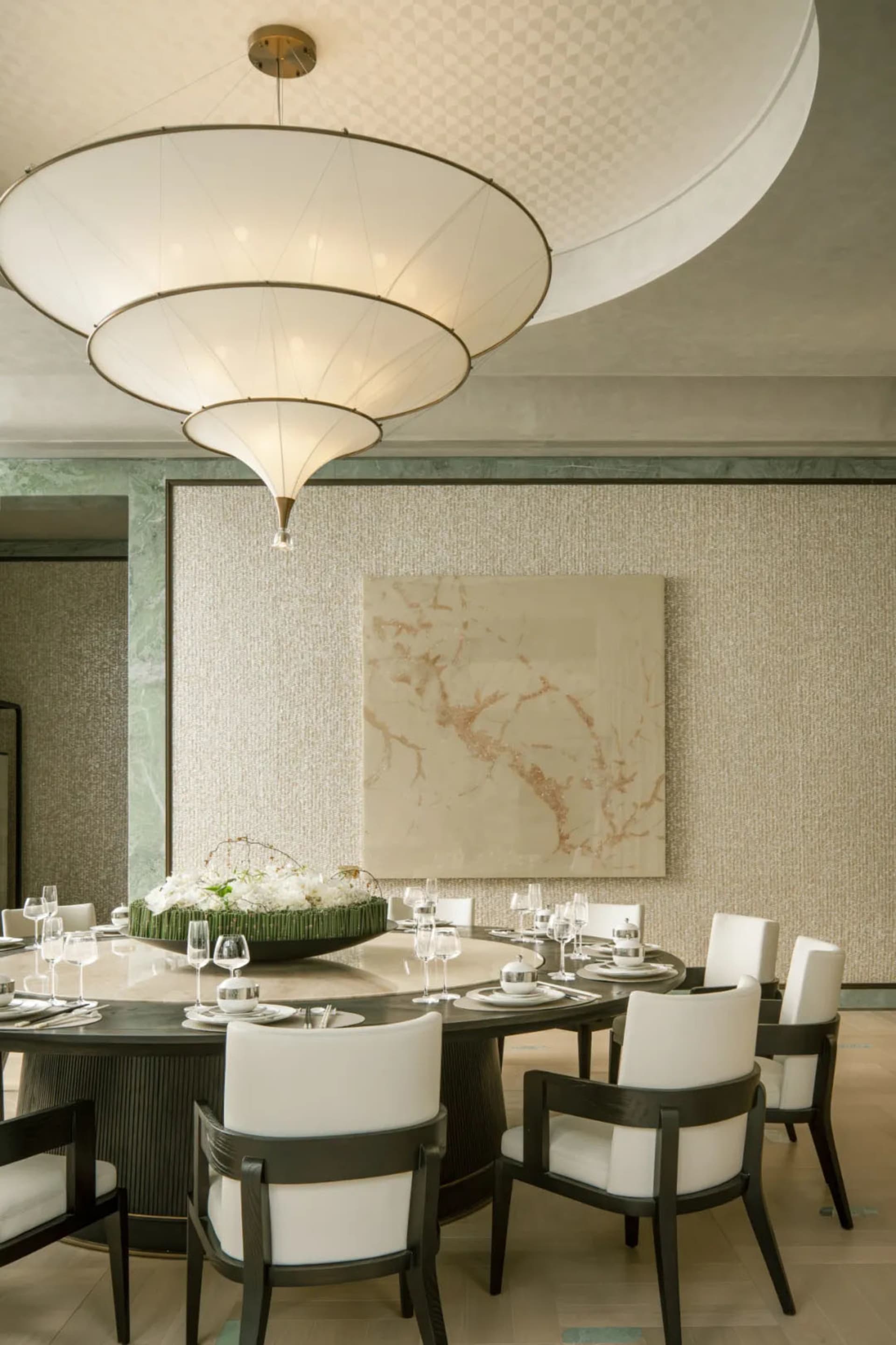 Dining room with a round table set for a meal, surrounded by chairs, a chandelier, and natural light from the windows