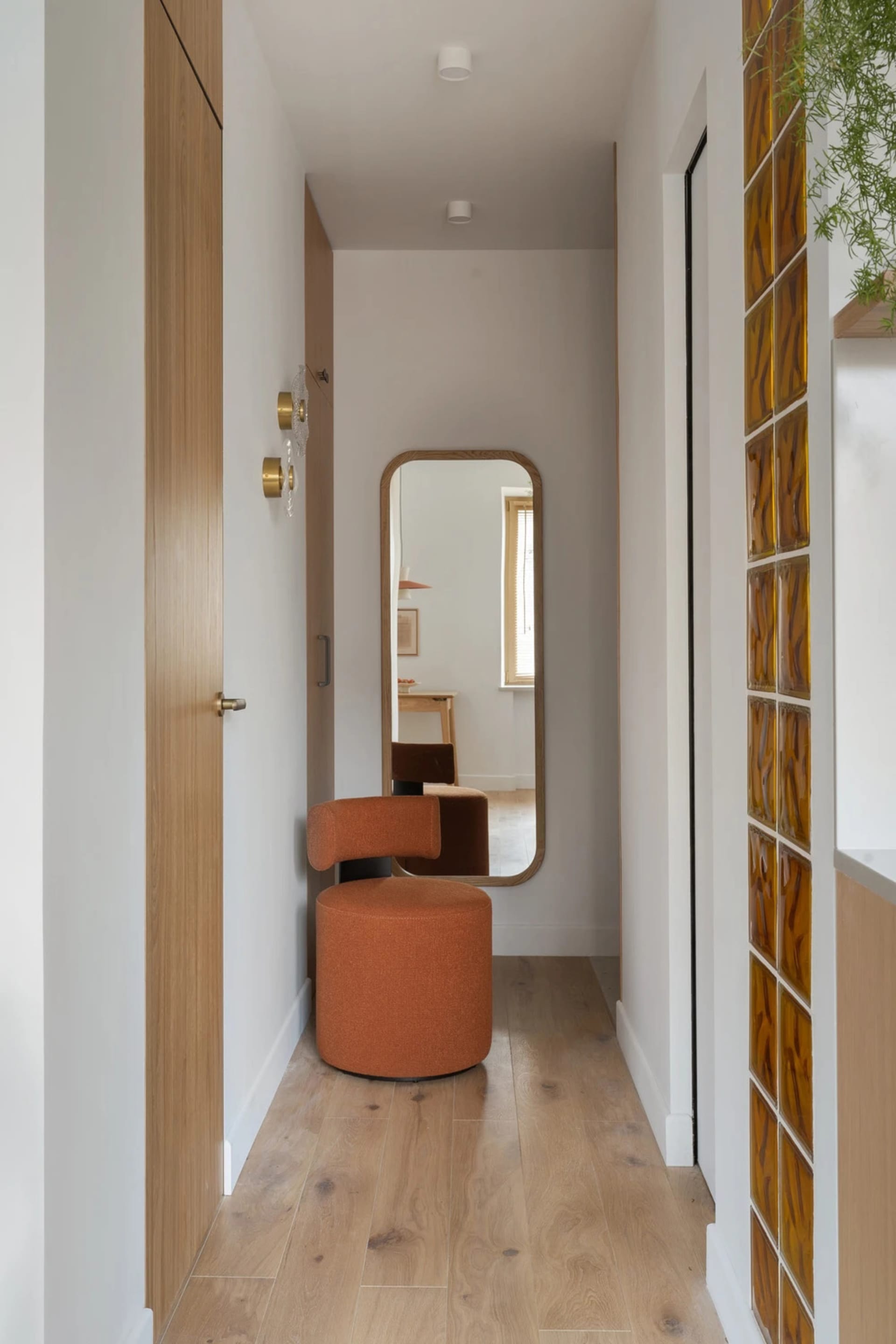 A modern hallway with a white wall, a brown wooden door, and various furniture pieces, including a chair, bench, and stool, under natural light