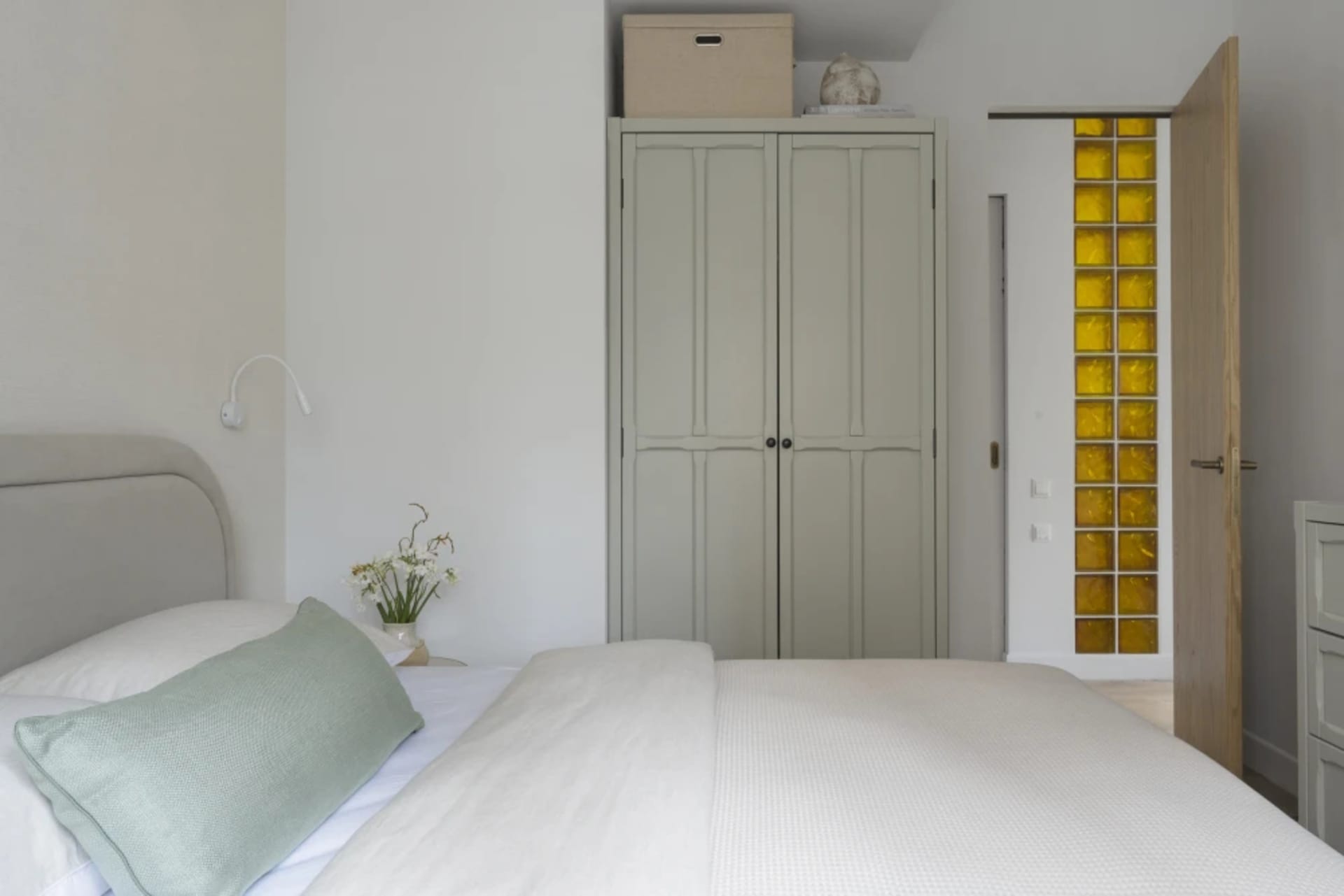 A minimalist bedroom with a bed, dresser, and closet, featuring a white comforter, two potted plants, and a clean, organized space