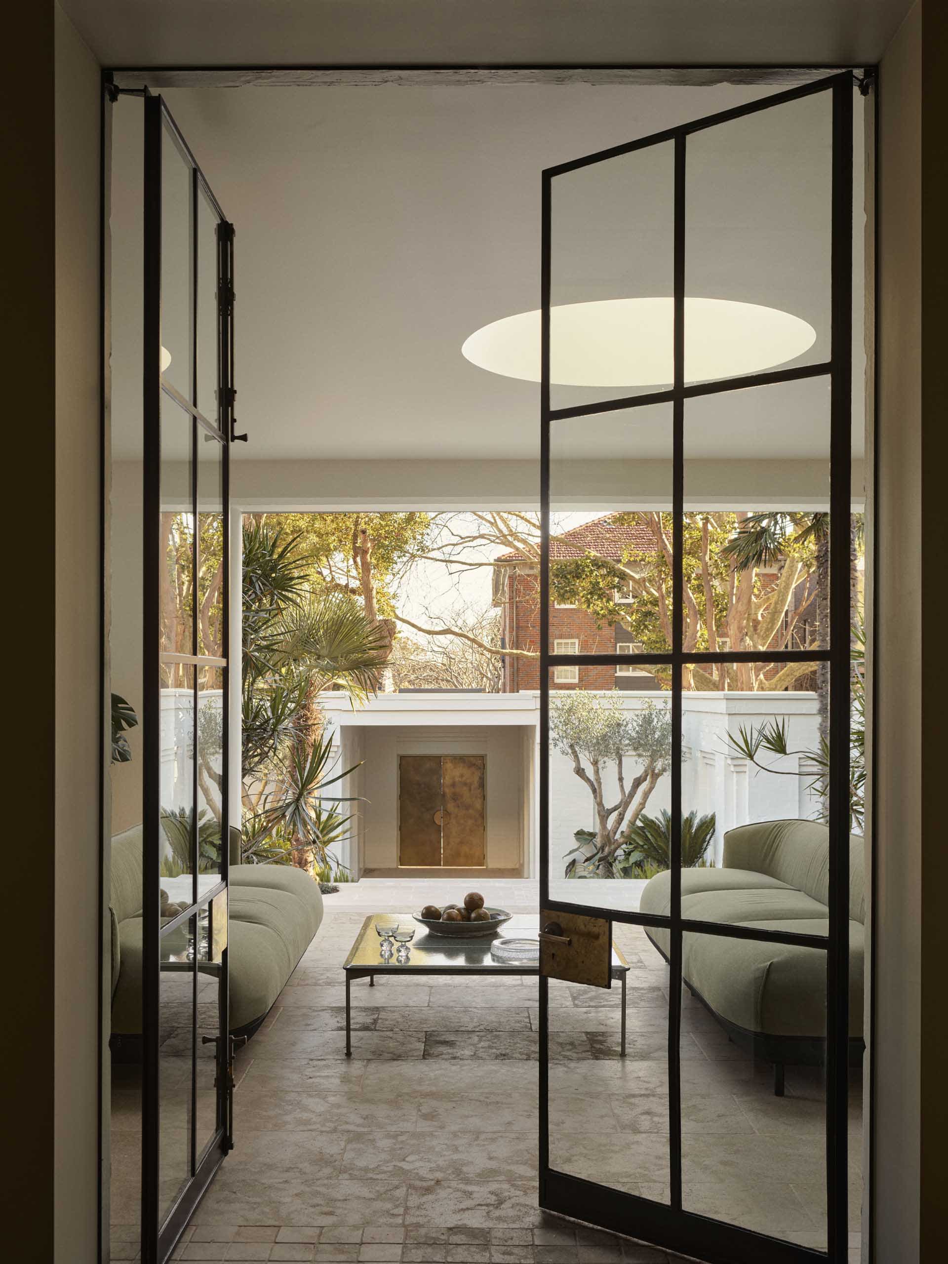 A modern living room with a glass door, a couch, a chair, and a dining table, surrounded by potted plants and natural light
