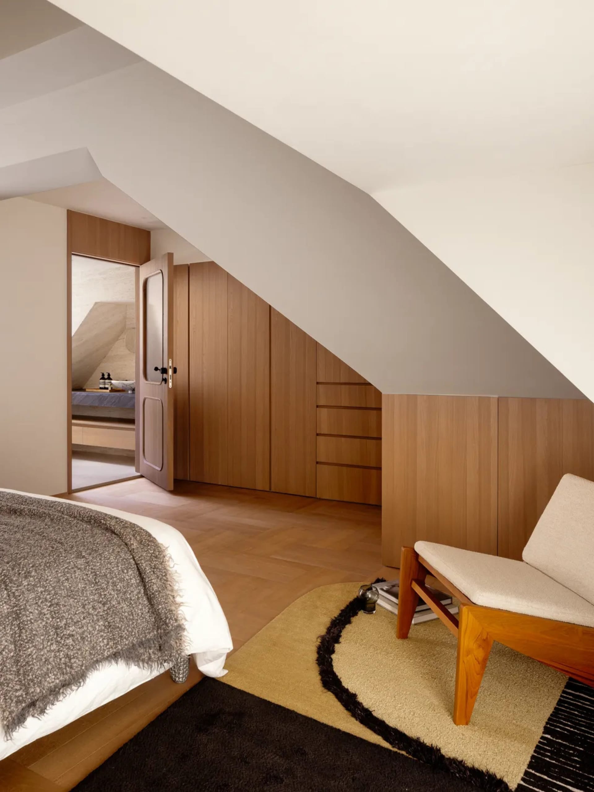 Cozy bedroom with a bed, chair, and couch, featuring wooden furniture and a rug, with natural lighting creating a warm atmosphere