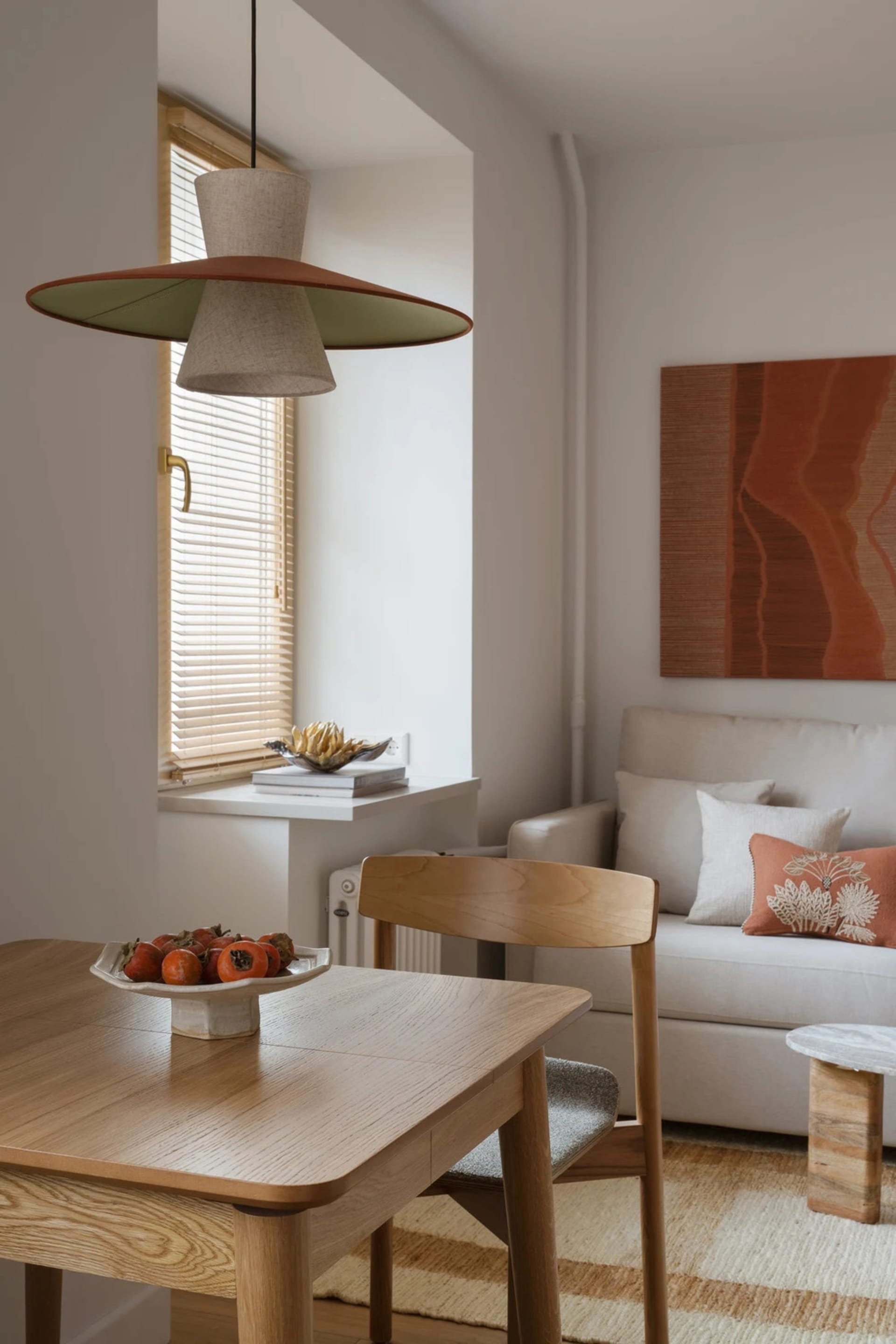 A modern living room with a wooden dining table, chairs, and couch, featuring a bowl of fruit and natural light from a window