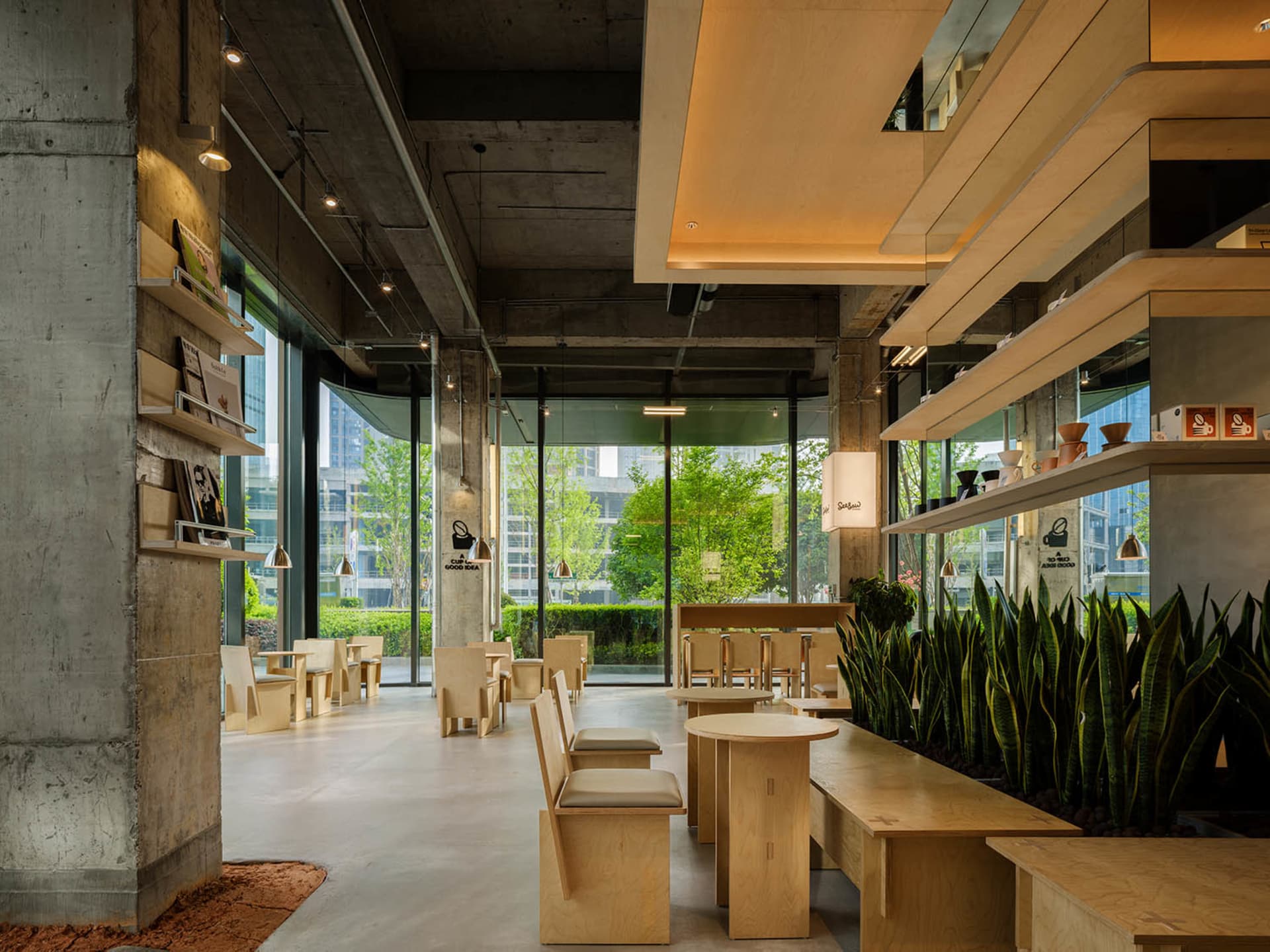 Modern restaurant interior with wooden furniture, green plants, and natural light from a large window, creating a bright and inviting atmosphere