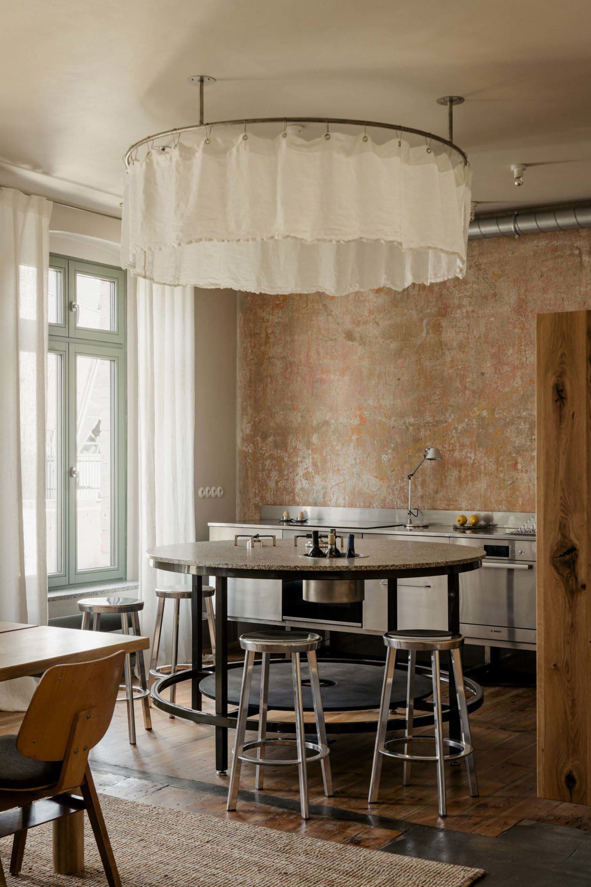 Residential kitchen with a wooden floor, white walls, and a dining table surrounded by chairs, with a sink and oven, in a dimly lit space