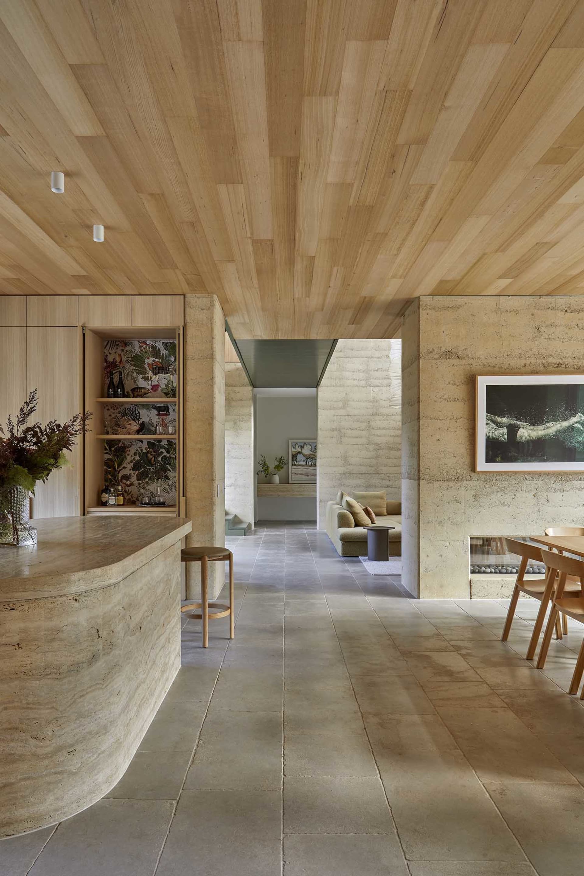Warm and inviting living room with a couch, chair, dining table, and kitchen area, decorated with plants, vases, and bottles, featuring wood and stone materials