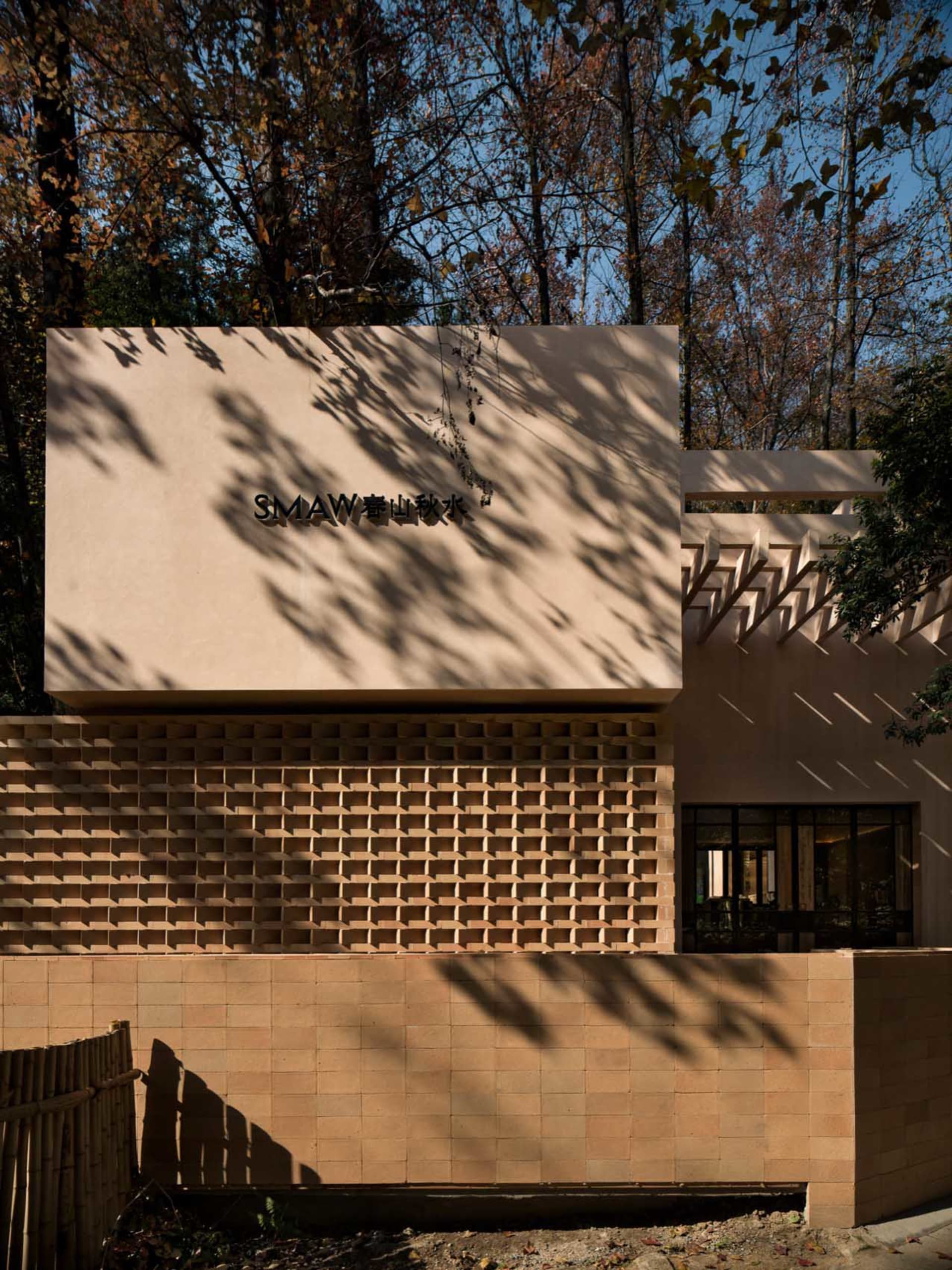 Photograph of a modern building with a tan exterior, featuring a large brick wall with a window and a door, set in a natural environment with trees and warm sunlight