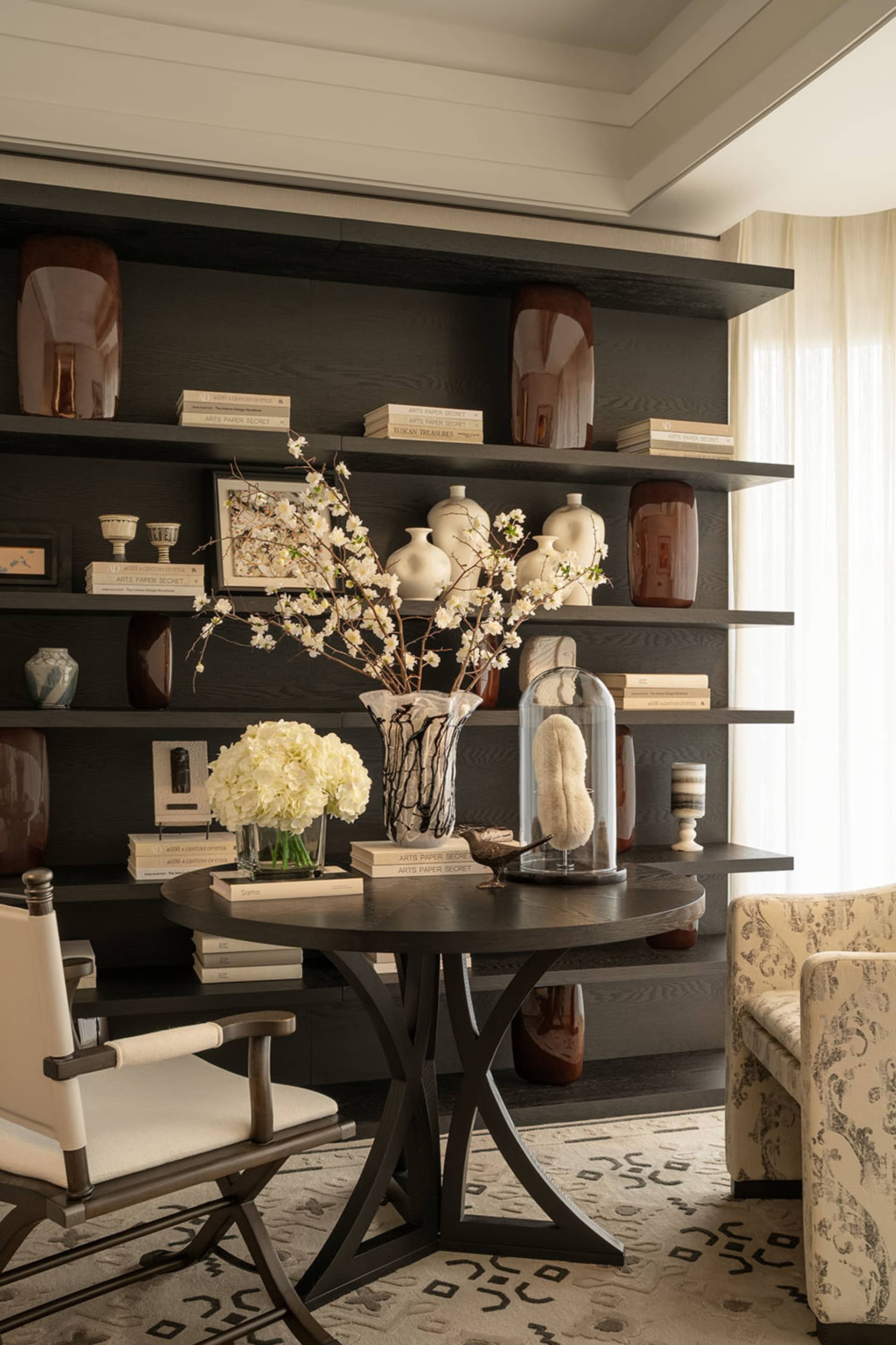 Cozy modern living space with a black table, vase of flowers, bookshelf, chair, couch, and natural light from windows
