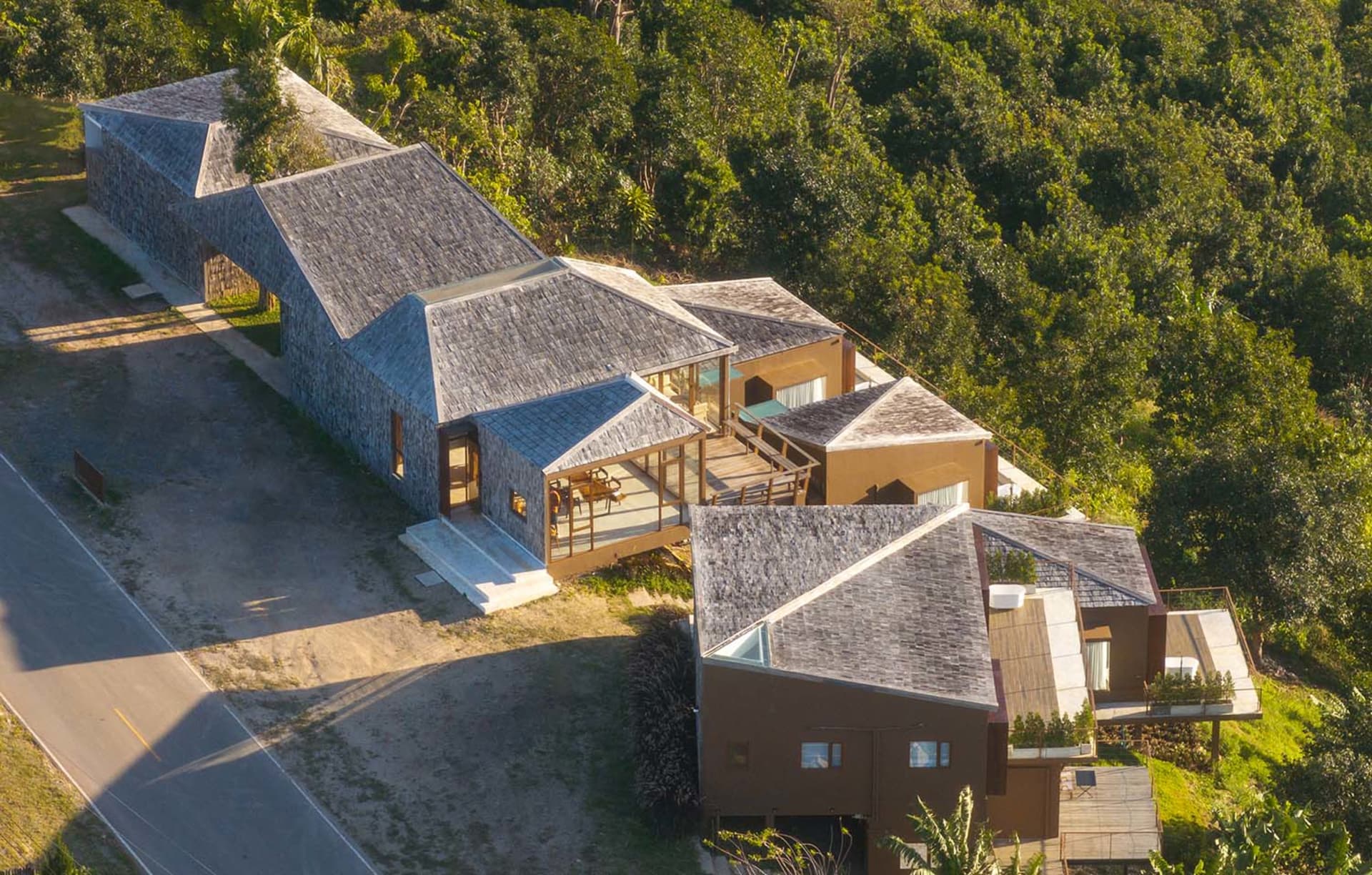 A modern house with a triangular roof, surrounded by trees, featuring a well-lit interior with a dining table and chairs, and a blend of natural materials and indoor-outdoor spaces