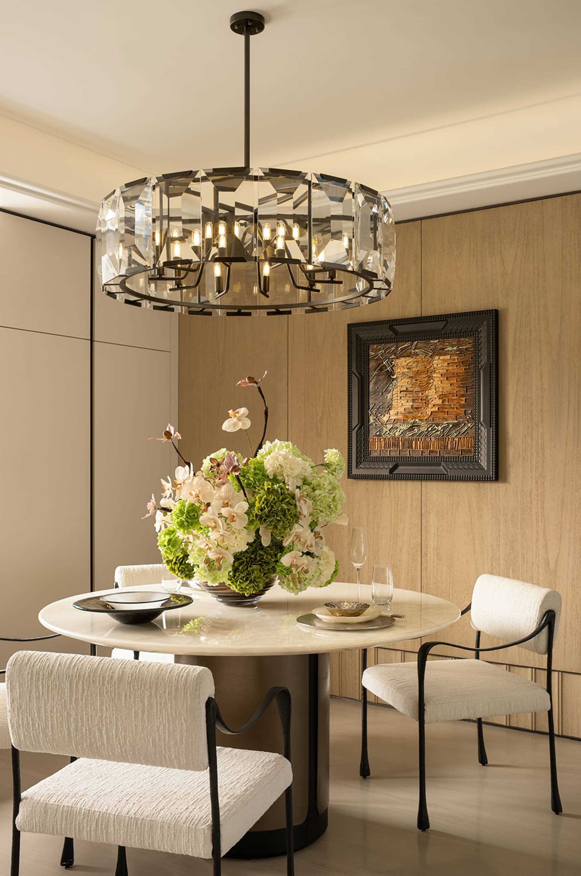Dining room with a wooden floor, a large dining table set with a vase, wine glasses, and a bowl, surrounded by chairs and a potted plant, under a hanging light fixture