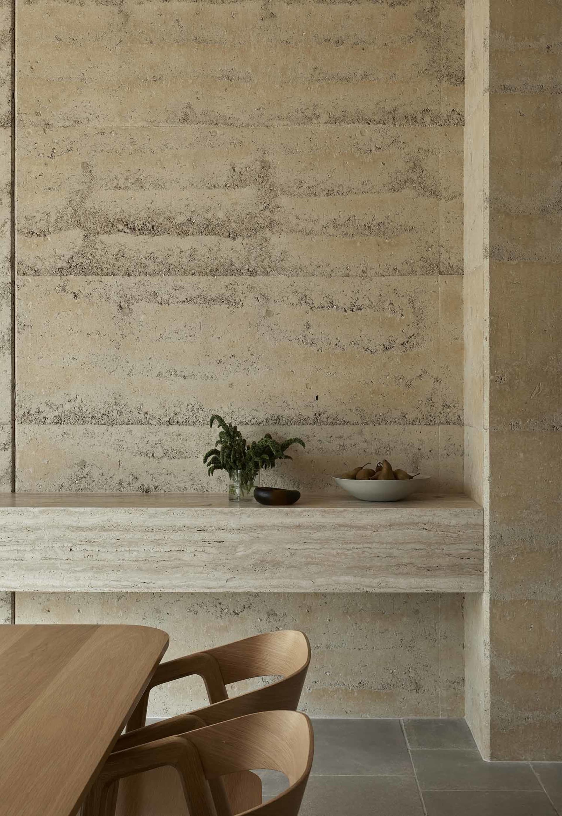 A modern dining area with a wooden table and chairs, surrounded by a stone wall and countertop, with a cozy dim lighting and a potted plant nearby