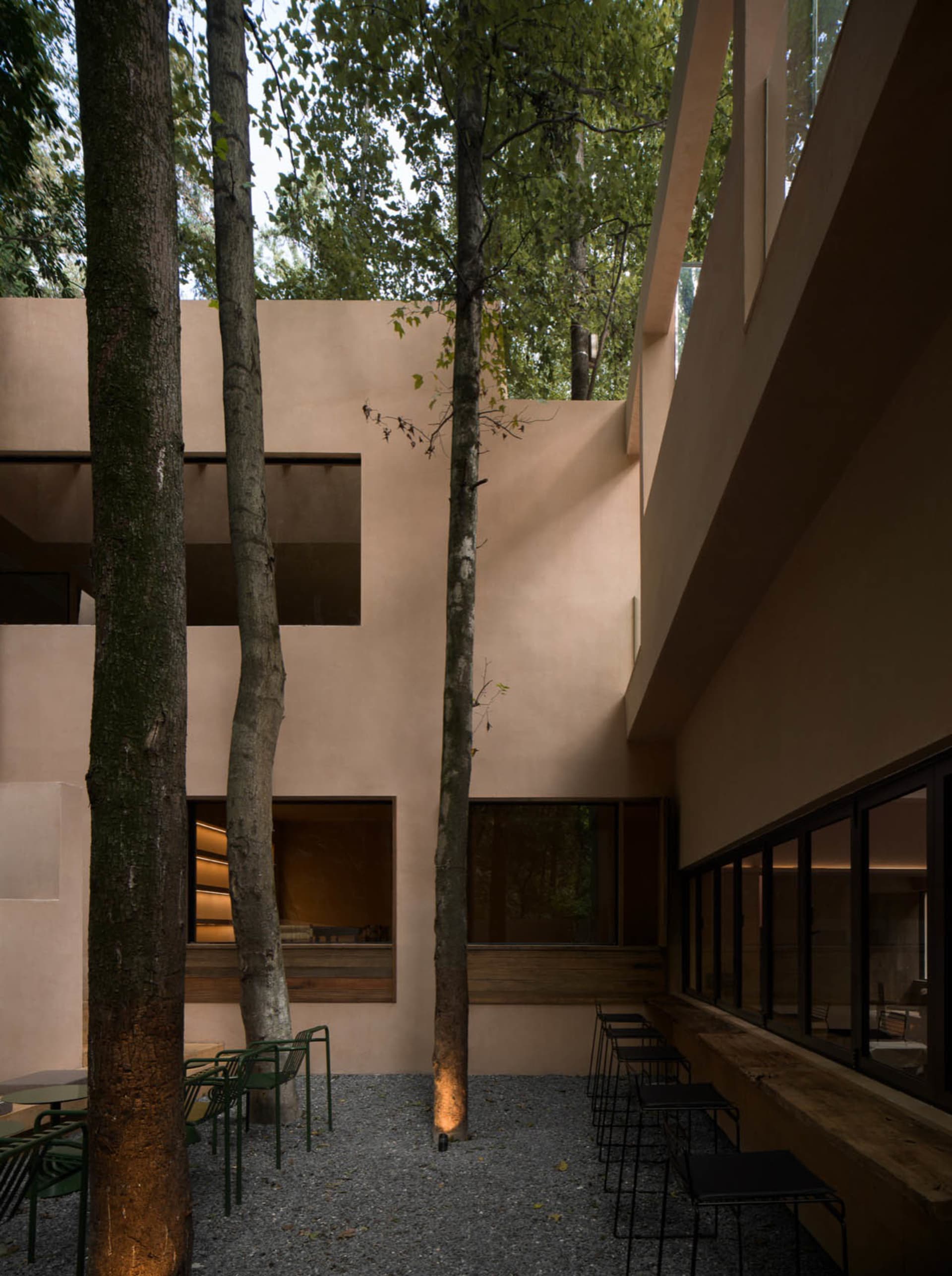 A modern building with a large patio area surrounded by trees, featuring a concrete exterior, a dining table, and chairs, with natural light and a relaxing atmosphere