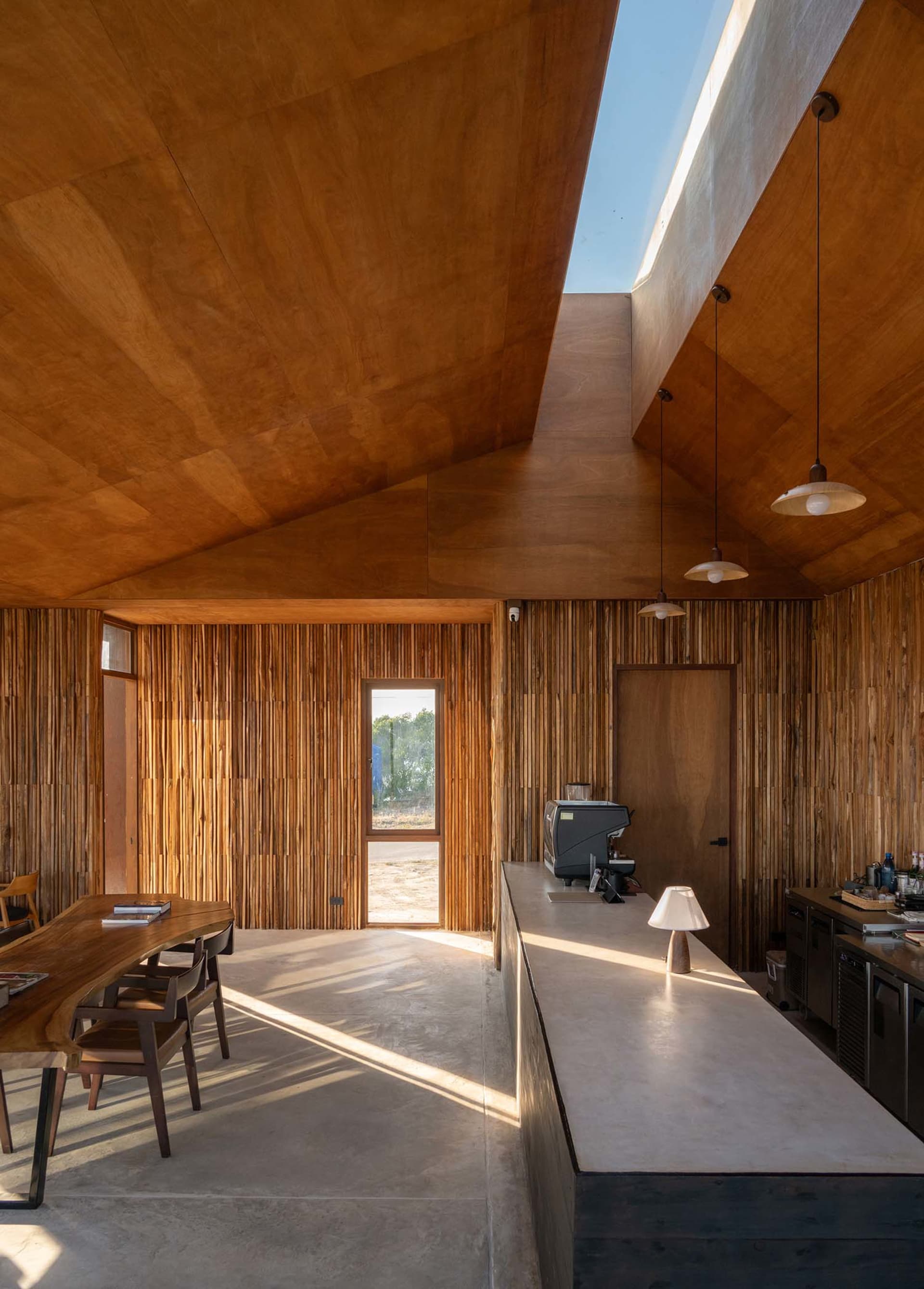 A modern dining room with a wooden floor, counter, and furniture, featuring a vaulted ceiling, skylight, and doorway, with soft and diffused lighting creating a warm and welcoming ambiance