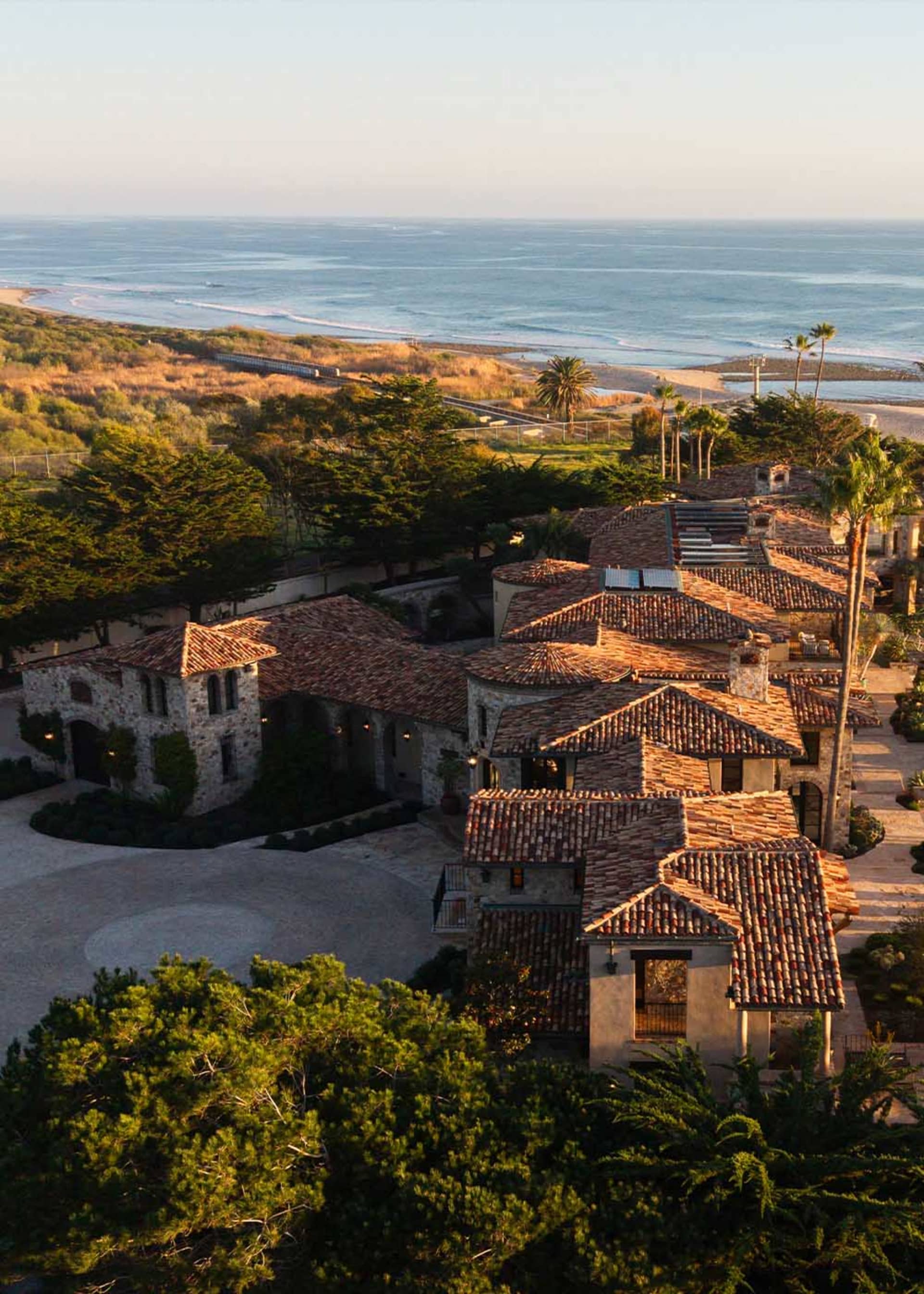 A large Spanish-style house with a red tile roof and various windows, situated on a hillside overlooking the ocean, bathed in natural light