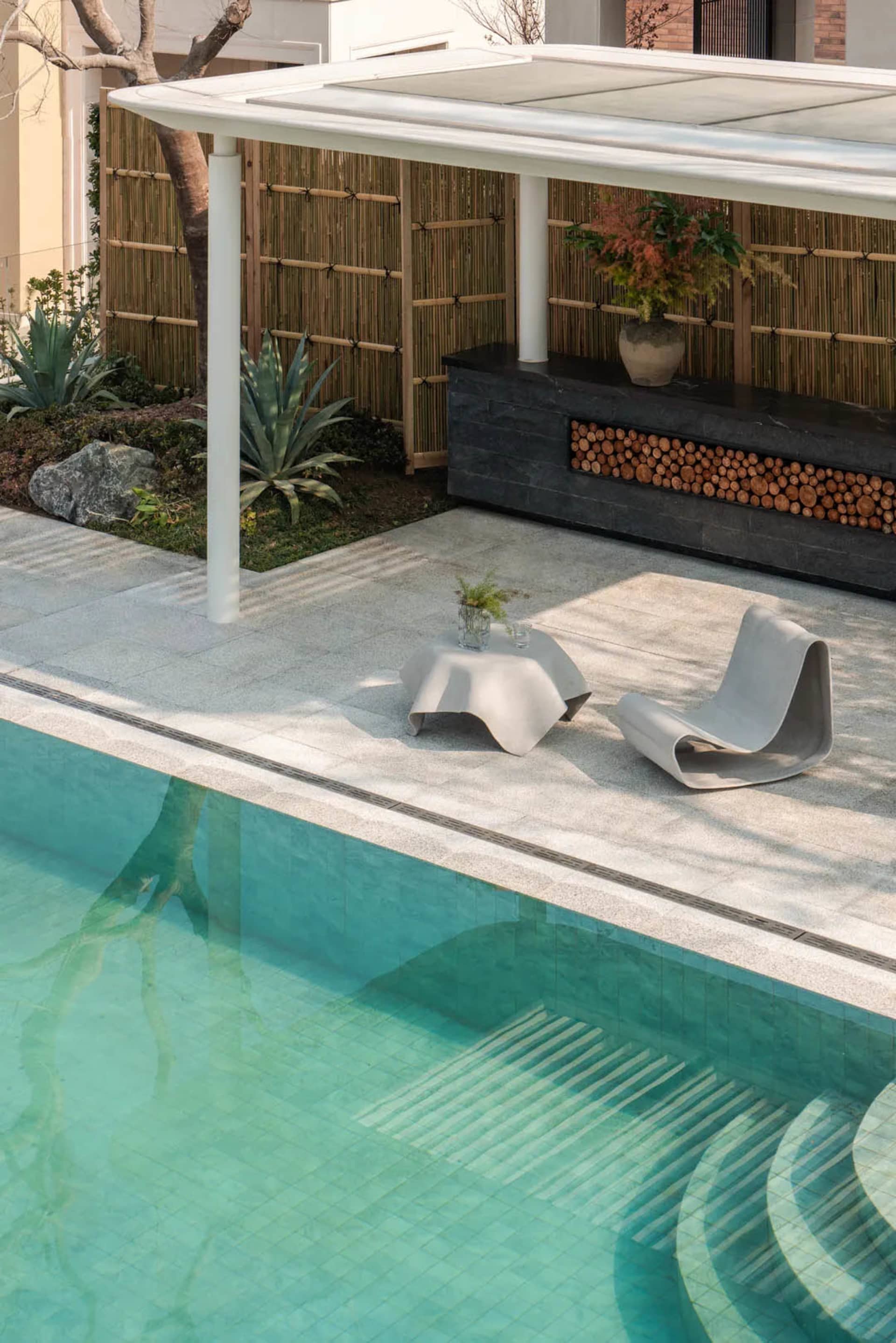 A serene outdoor patio area with a pool, wooden fence, and potted plants, featuring a bench and chair for relaxation