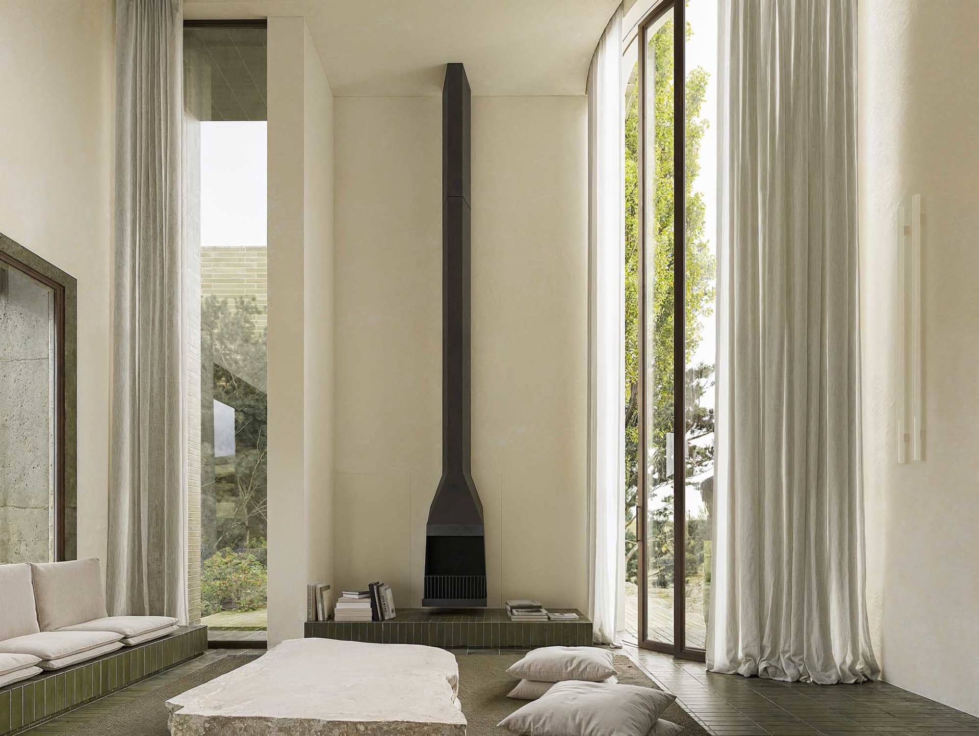Cozy modern living space with a stone fireplace, a large window, and natural materials, featuring a bed, couch, chair, and books