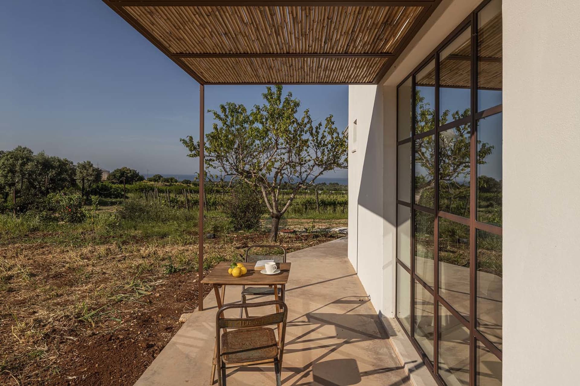 A modern outdoor dining space with a table, chairs, and a shade structure with a woven roof, surrounded by a fence in a residential coastal area