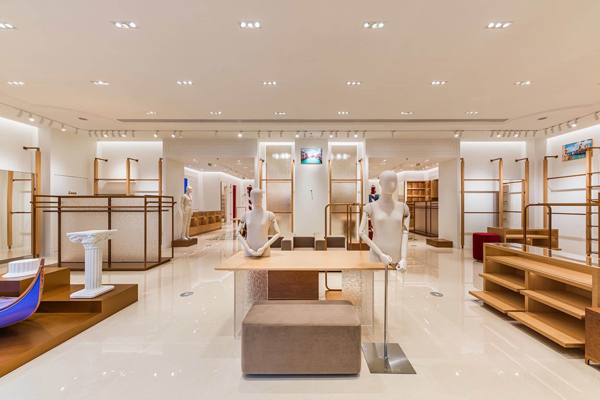 Spacious luxury retail showroom interior with light oak clothing display racks, white mannequins, polished marble floor, and recessed ceiling lighting.