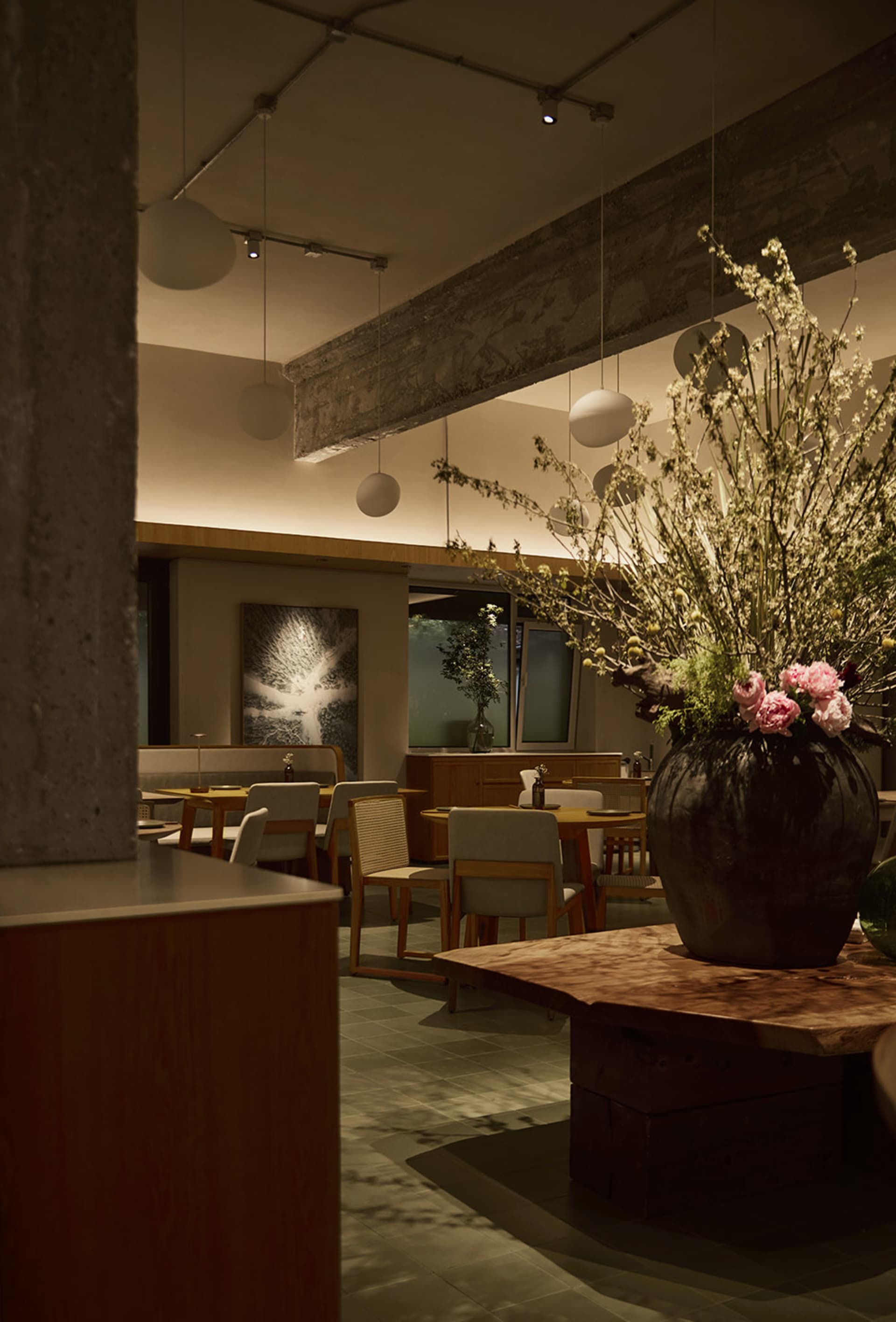 Interior view of a modern wabi-sabi style restaurant featuring wooden tables, upholstered chairs, exposed concrete elements, and a large floral centerpiece in a dark ceramic vase.