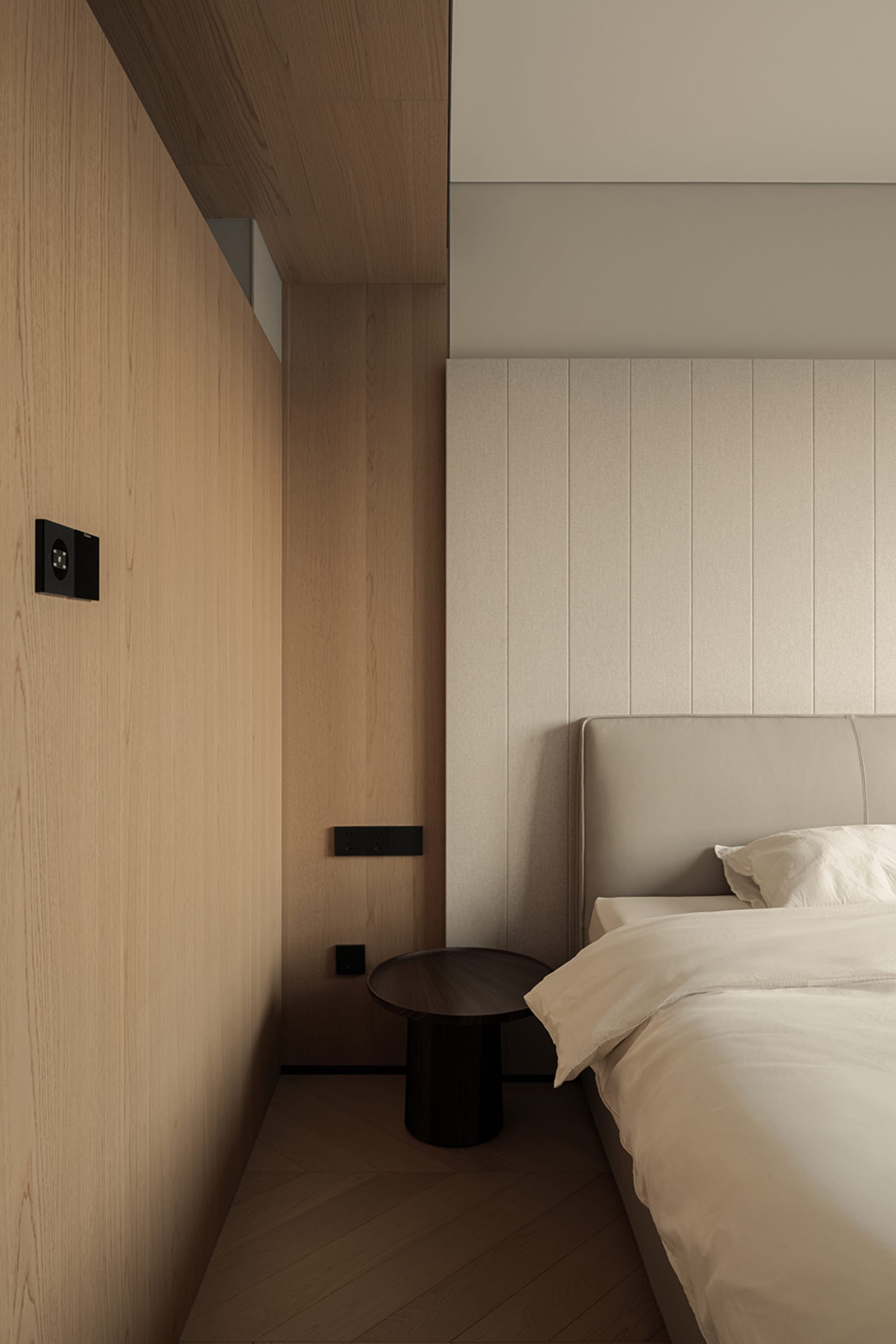 Modern minimalist bedroom featuring vertical light oak wood paneling on the left wall, a beige upholstered headboard with matching fabric wall paneling, a dark round wooden bedside table, and crisp white bedding.