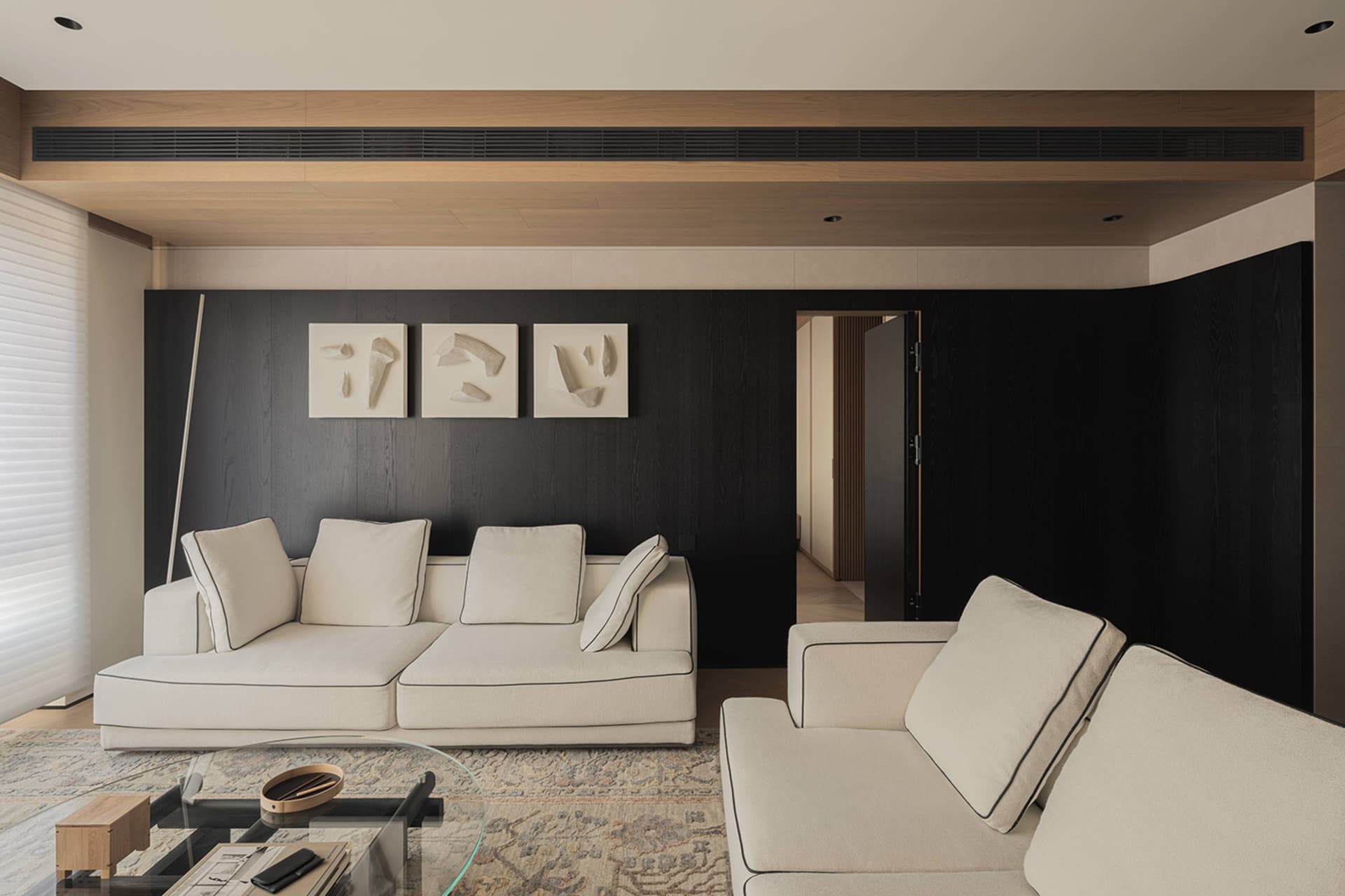 Modern living room featuring a dark wood accent wall with three white abstract artworks, large off-white L-shaped sectional sofa, glass coffee table on a patterned area rug, and wooden ceiling detail.
