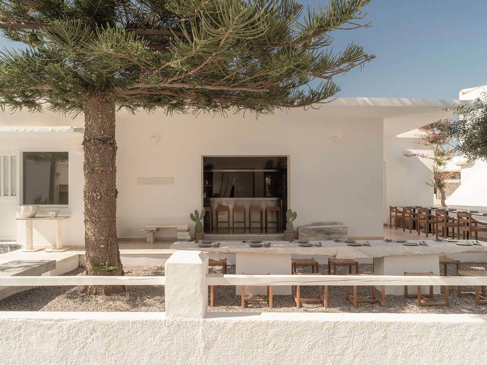 Outdoor dining courtyard with long stone table, wooden stools, whitewashed building, and a large pine tree under clear blue sky