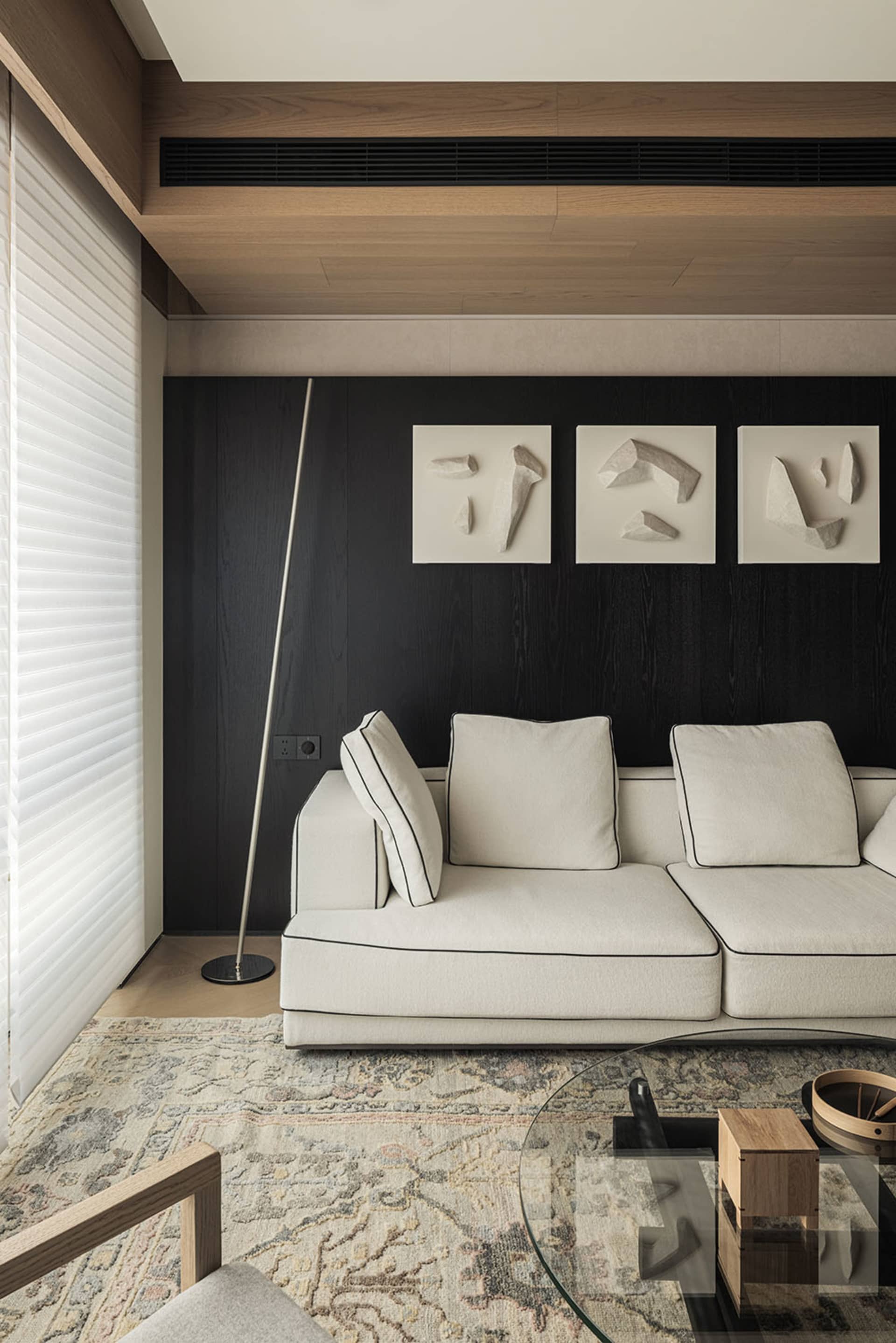 Modern living room featuring a white sectional sofa, dark wood accent wall with three framed sculptural artworks, patterned area rug, and wooden ceiling details.