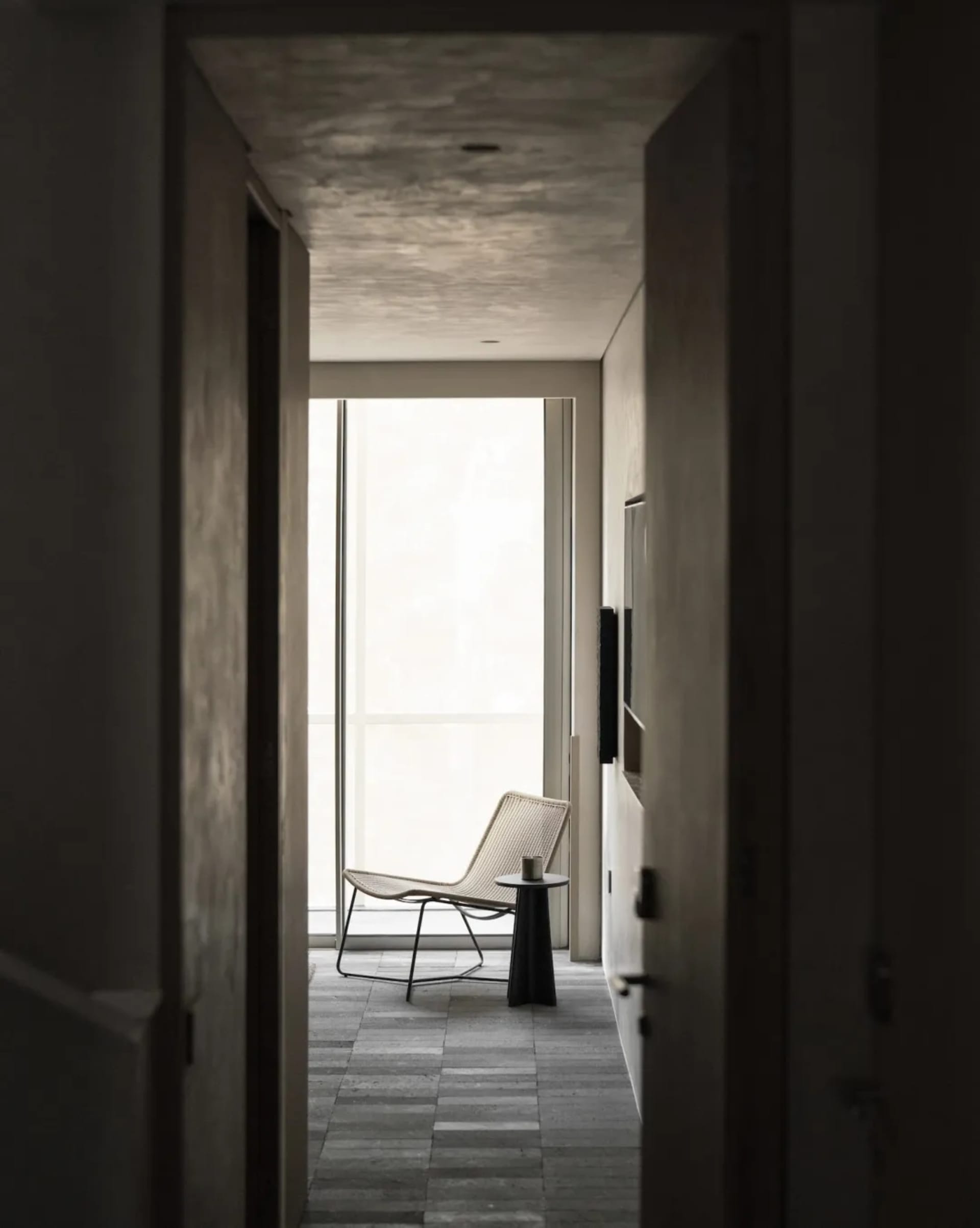 View through a doorway into a minimalist concrete hallway featuring a woven lounge chair, black side table, and large window with natural light.
