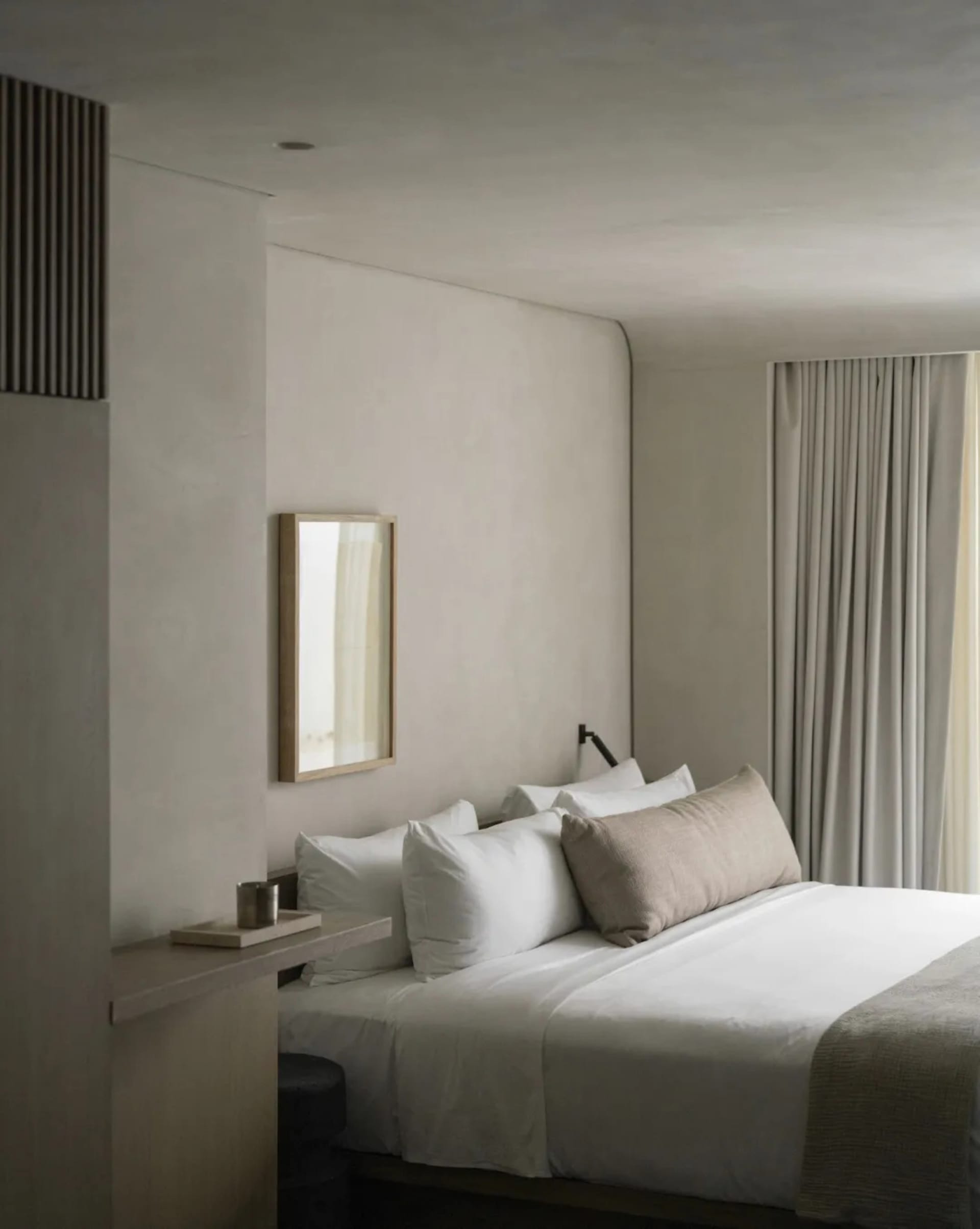 Modern minimalist bedroom with textured plaster walls, white bedding, beige linen pillows, wooden-framed mirror, and soft natural light from sheer curtains.