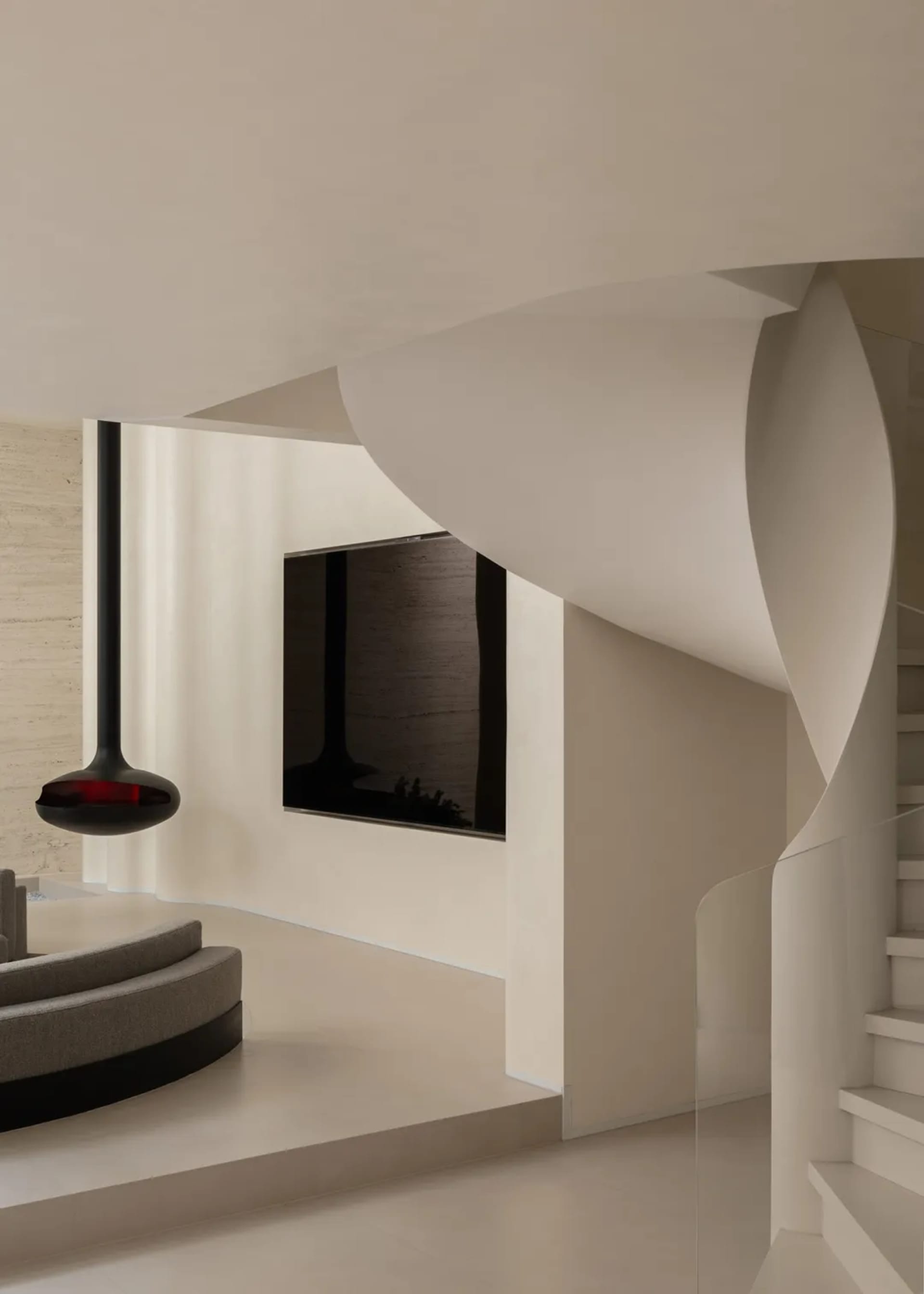 Modern minimalist interior featuring a sculptural curved white staircase, floating black pendant light, built-in TV, and curved grey sofa against textured concrete walls.