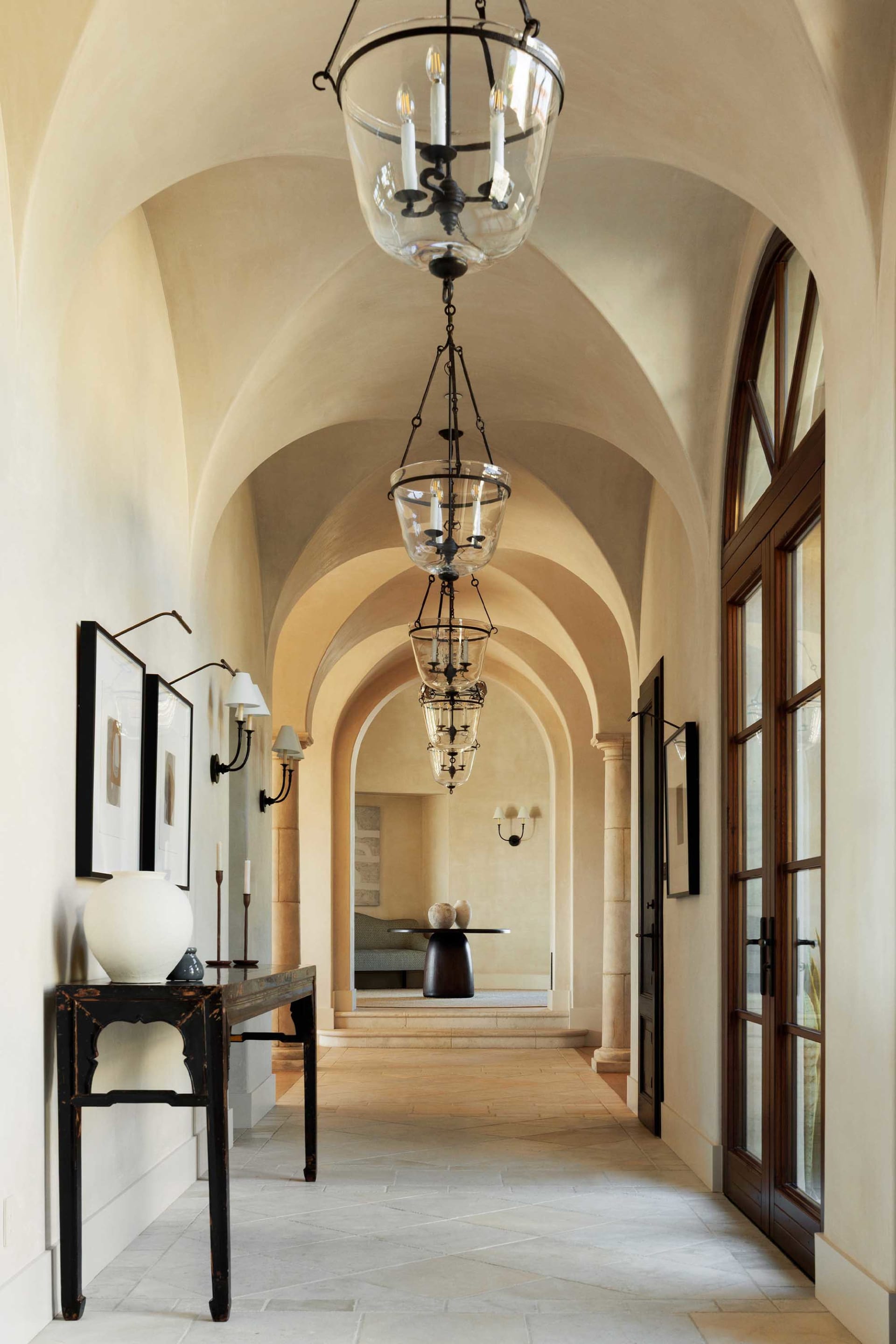 Long arched hallway in Tuscan style featuring multiple glass and iron lanterns, a distressed black console table with white ceramic vase, framed artwork, and limestone flooring leading to a seating area.