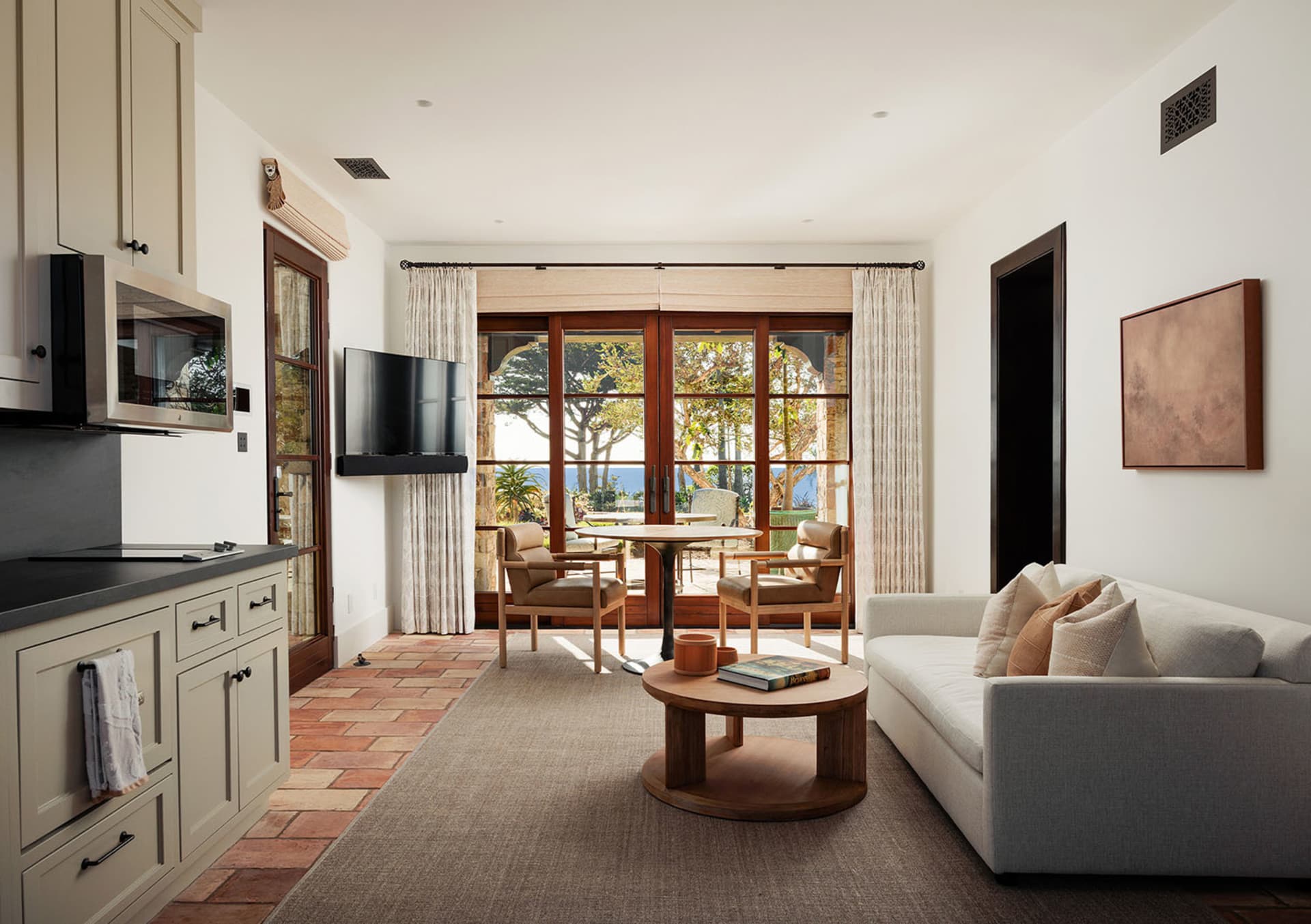 Bright open-plan living room featuring terracotta floor tiles, a light gray sofa, round oak coffee table, two leather armchairs, wall-mounted TV, and large glass doors opening to a coastal terrace with ocean view.