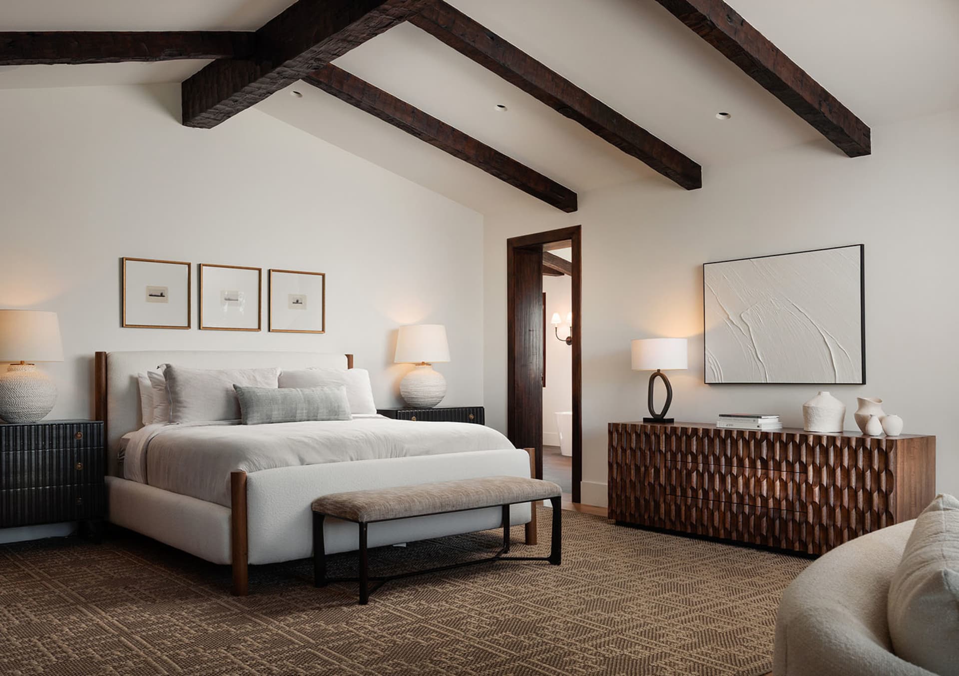 Modern rustic bedroom with white walls, exposed dark wooden ceiling beams, upholstered bed with neutral linens, textured wooden console, and herringbone carpet.