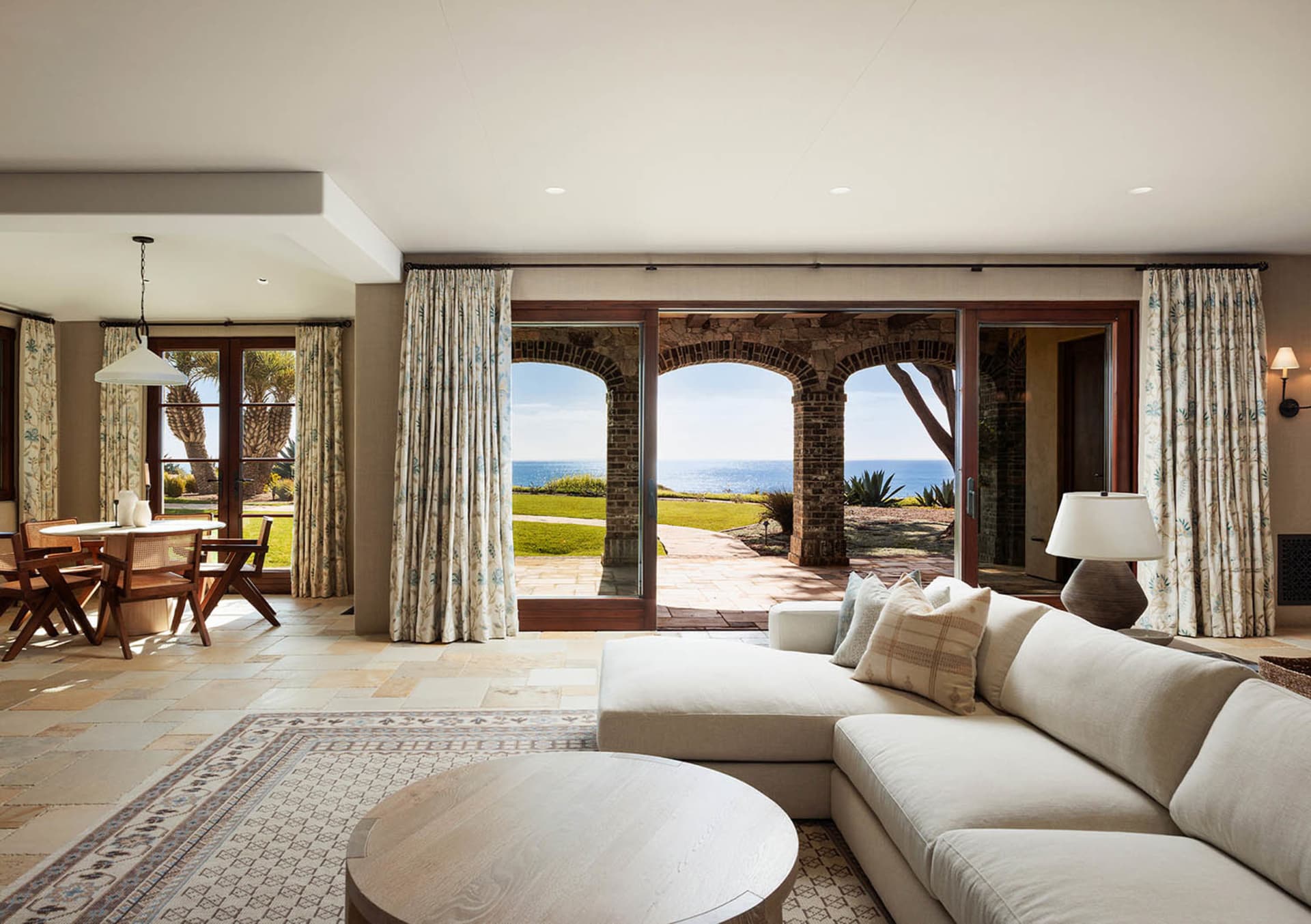 Bright coastal living room featuring a large L-shaped sectional sofa, round wooden coffee table, patterned area rug, and expansive ocean view through three brick arches leading to a patio.