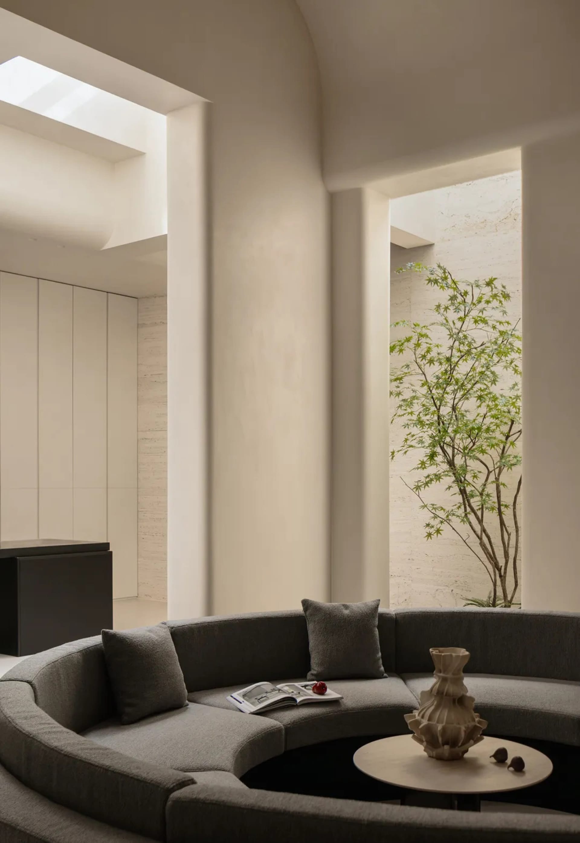 Modern minimalist living room featuring a large curved grey sectional sofa, round coffee table with sculptural vase, travertine walls, and a small tree in an open courtyard under a skylight.