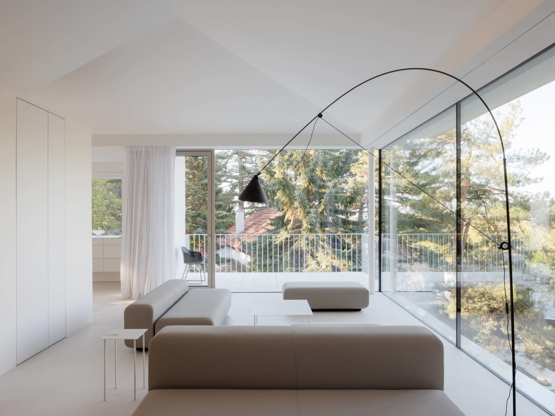 Modern minimalist living room featuring low-profile beige fabric sofas, a large black arched floor lamp, white walls, and floor-to-ceiling glass windows overlooking a lush green forest and balcony.
