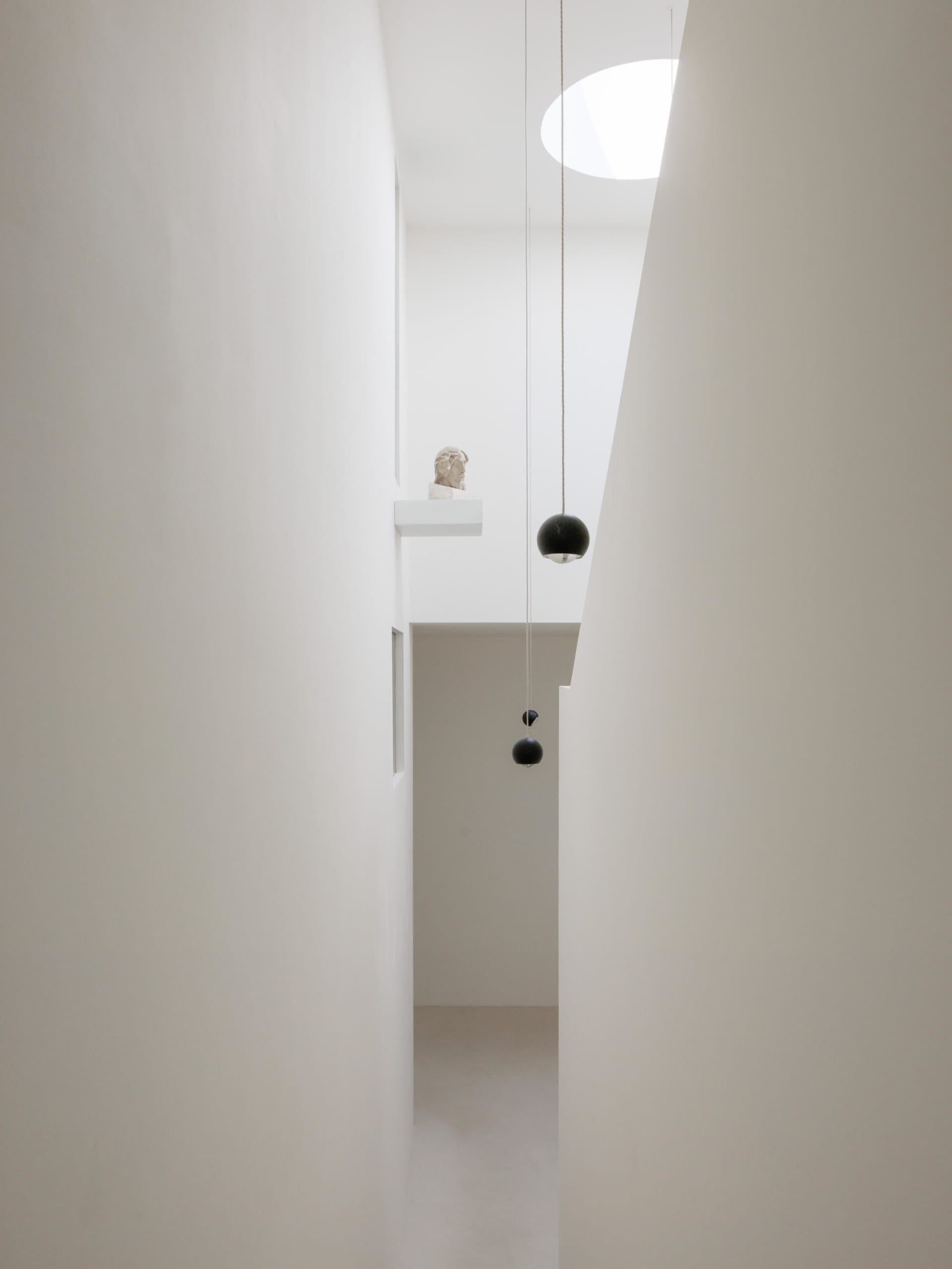 View looking up into a minimalist white double-height hallway featuring two black spherical pendant lights, a classical bust on a high ledge, and a large circular skylight.