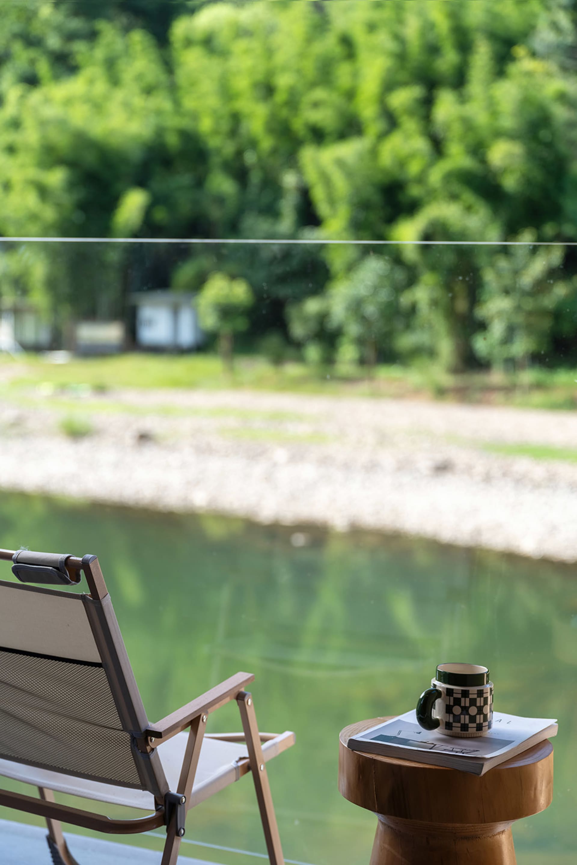 Wooden folding chair and teak side table with coffee mug and magazine, positioned by a large window overlooking a green river and forested hills.
