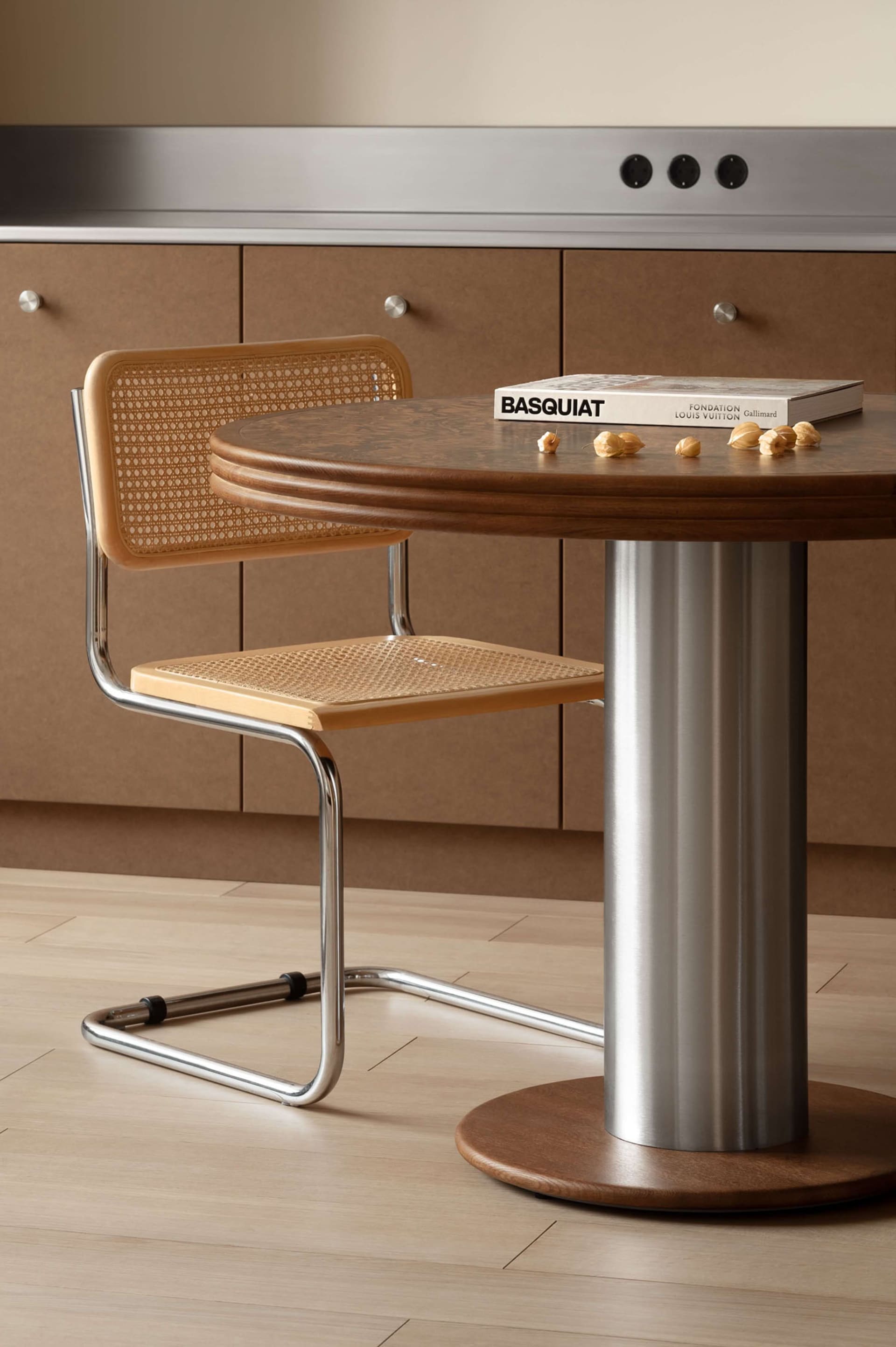 A light wood cane chair with chrome frame sits beside a round walnut pedestal dining table featuring a stainless steel column base, with a Basquiat book and walnuts on the tabletop.