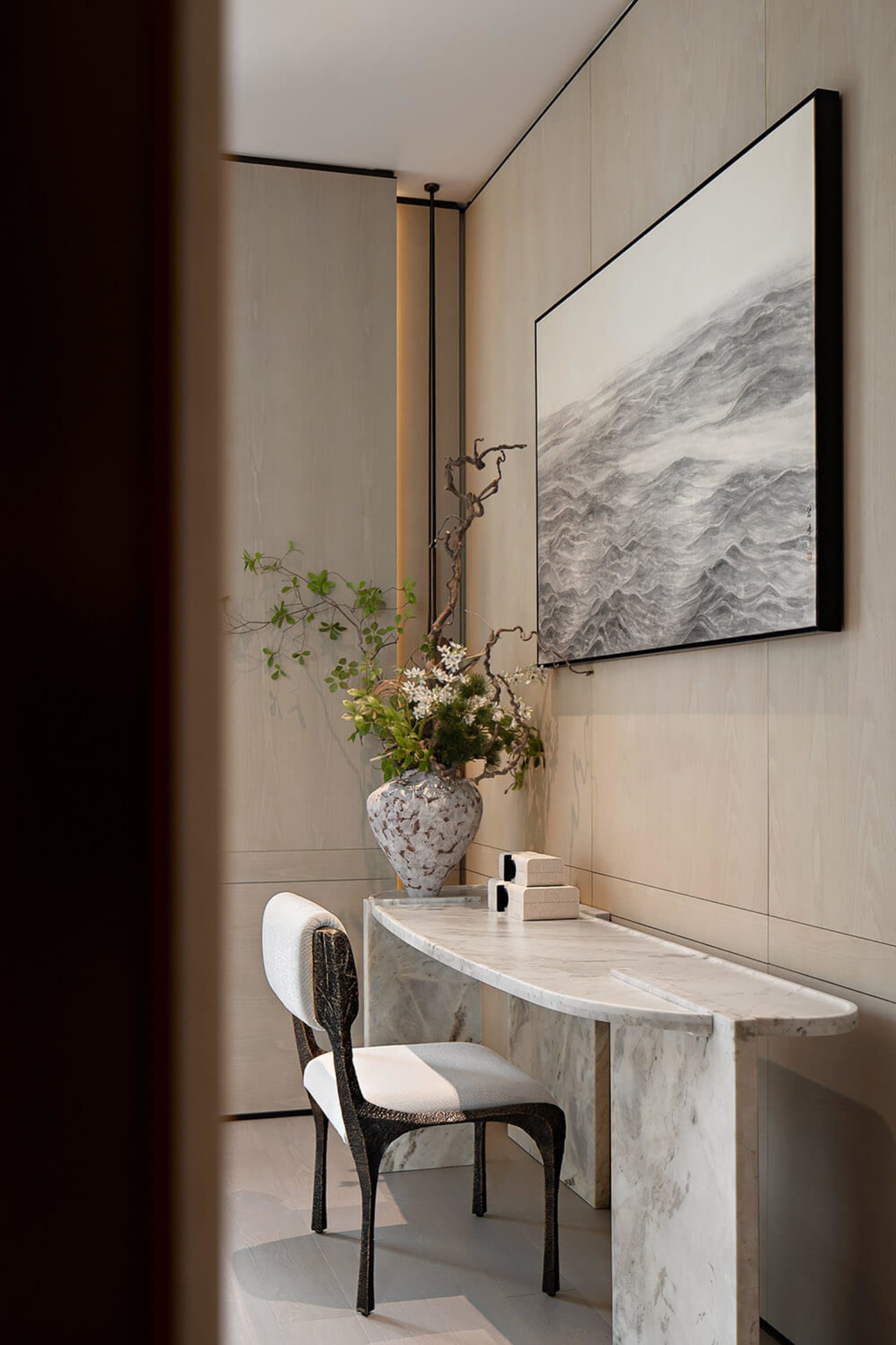 Minimalist entryway featuring a curved white marble console table, a dark sculptural chair, a large ceramic vase with floral arrangement, and a black-framed ink wash painting of waves on a light wood-paneled wall.