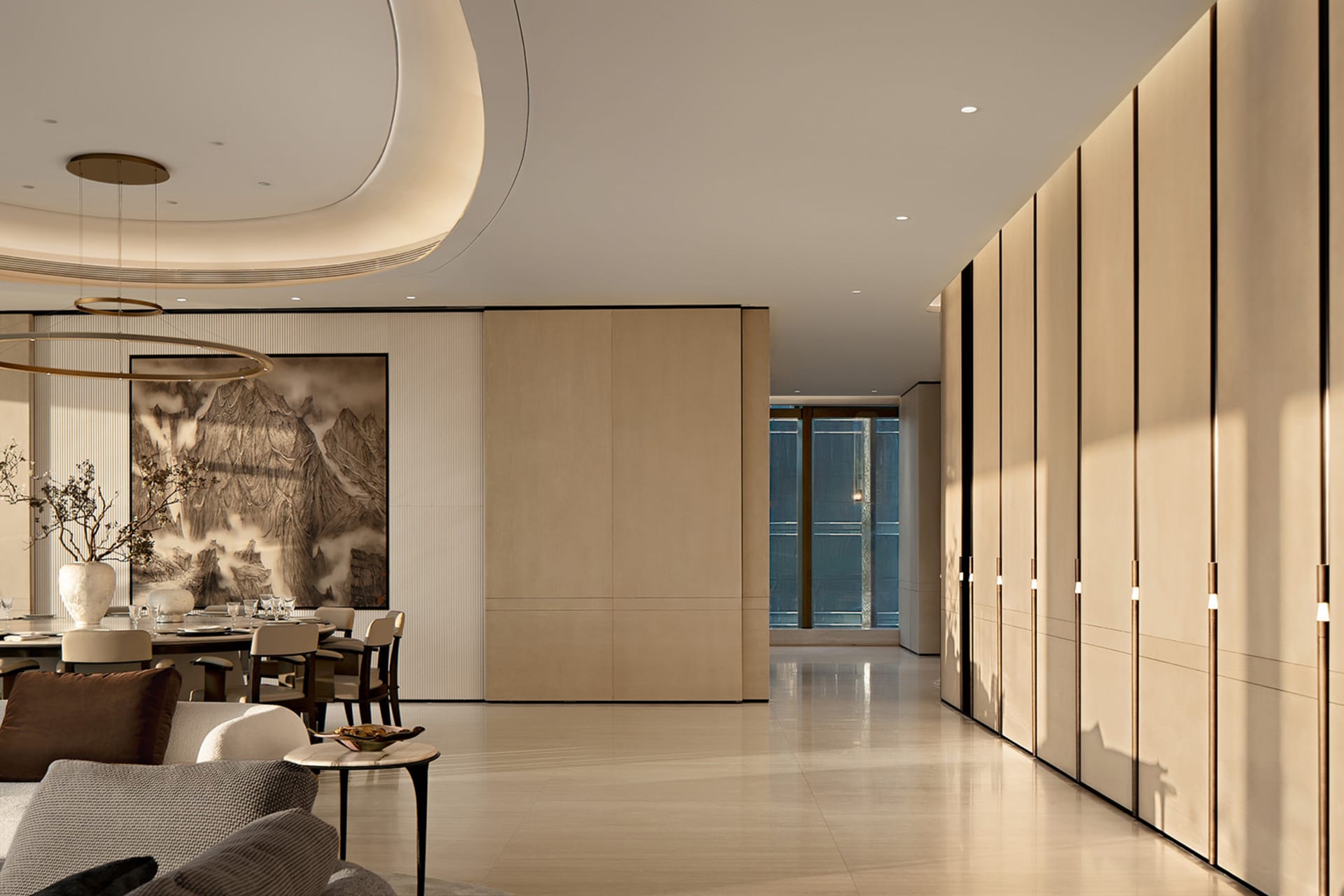 Modern open-plan dining and living space featuring a large circular chandelier, neutral upholstered seating, a round dining table, and a prominent black-and-white mountain ink painting on a vertically ribbed wall, complemented by warm oak paneling and a glossy light floor.