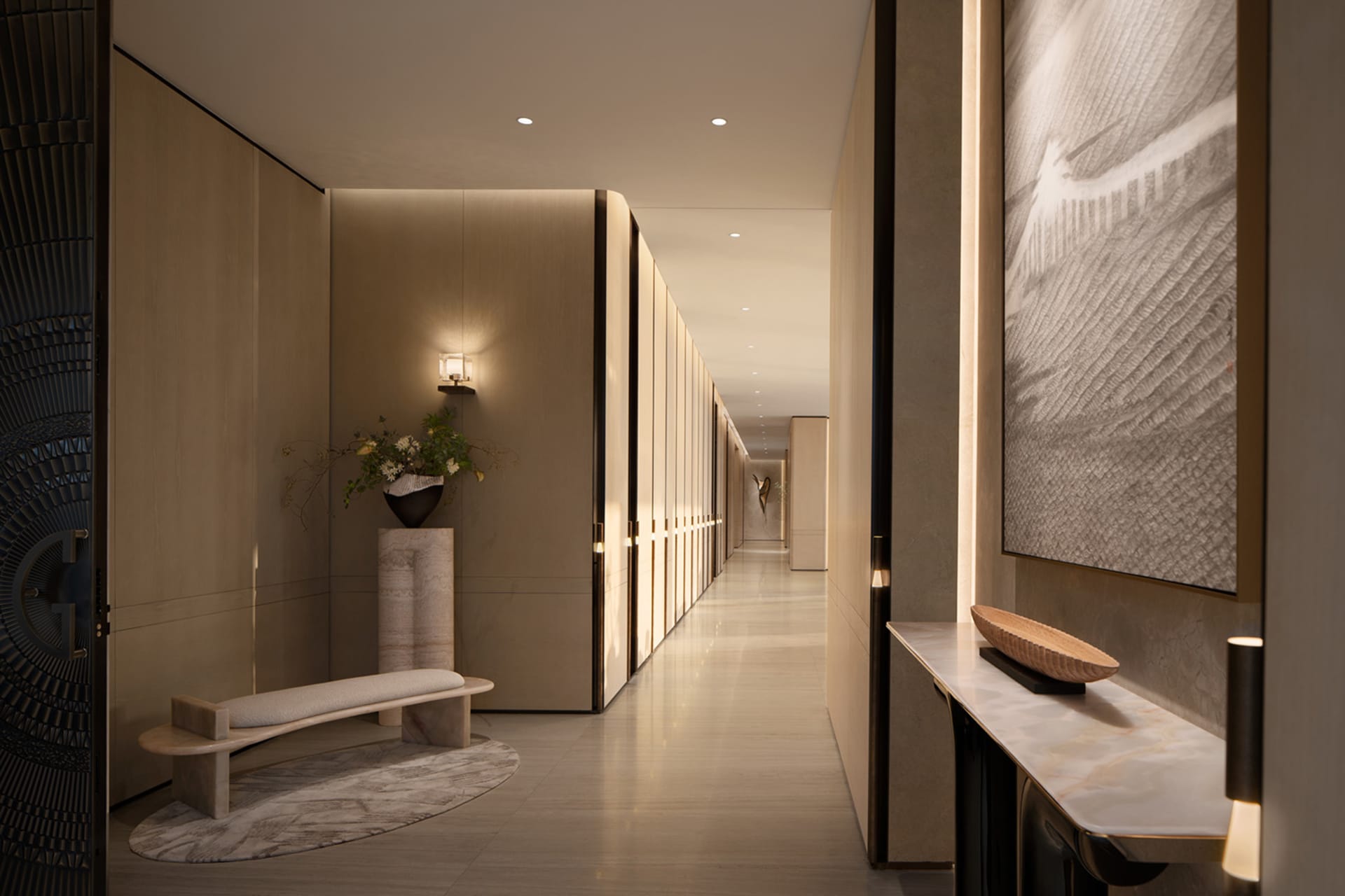 Modern minimalist luxury hallway featuring a travertine console table, wall sconces, large abstract artwork, and a curved stone bench with floral arrangement on a marble pedestal.
