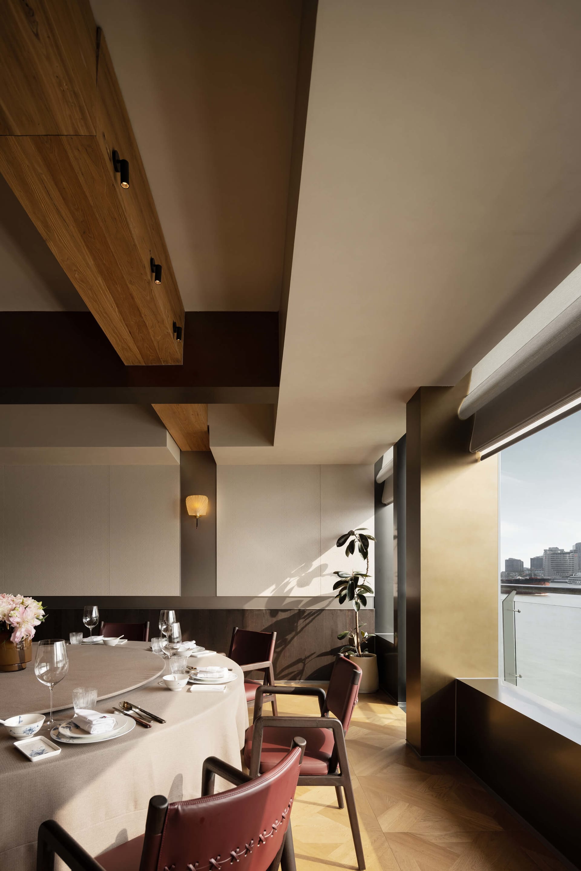 Modern elegant dining room featuring a large round table with beige tablecloth, burgundy leather chairs, walnut wood ceiling beams, and a large window with harbor view.