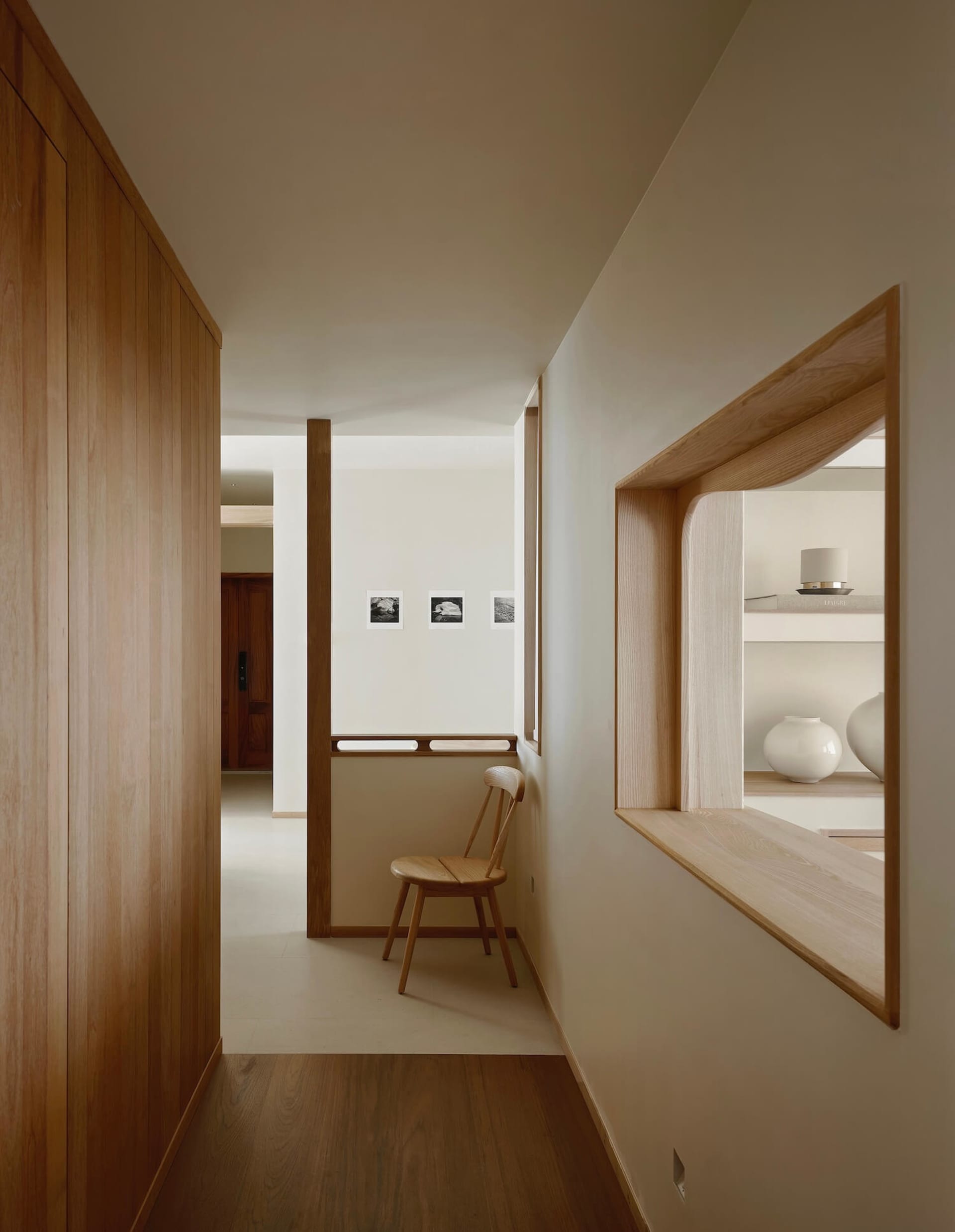 Modern minimalist hallway featuring light oak wood paneling, a built-in curved display niche with white ceramic vases, and a wooden chair against a white wall with small framed photographs.