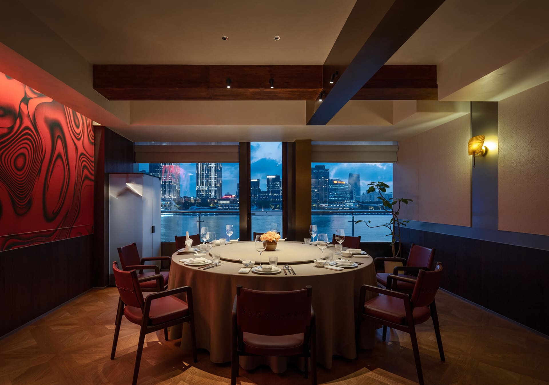 Elegant private dining room featuring a large round table with red leather chairs, dramatic red marble wall, dark wood beams, and large windows showing a twilight harbor city view.