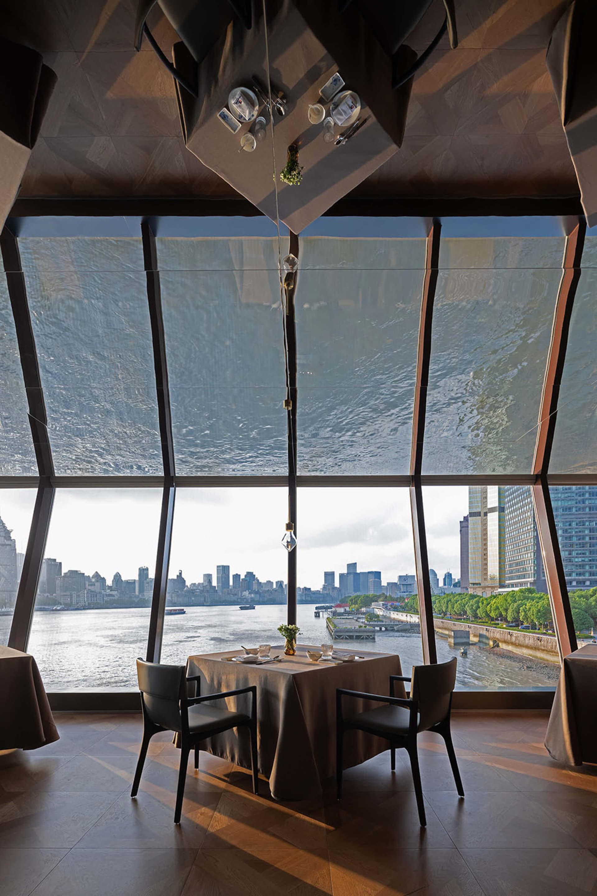 Modern fine-dining table with black leather chairs positioned in front of enormous angled windows revealing a river and dense city skyline, under a striking geometric wood-paneled ceiling feature.